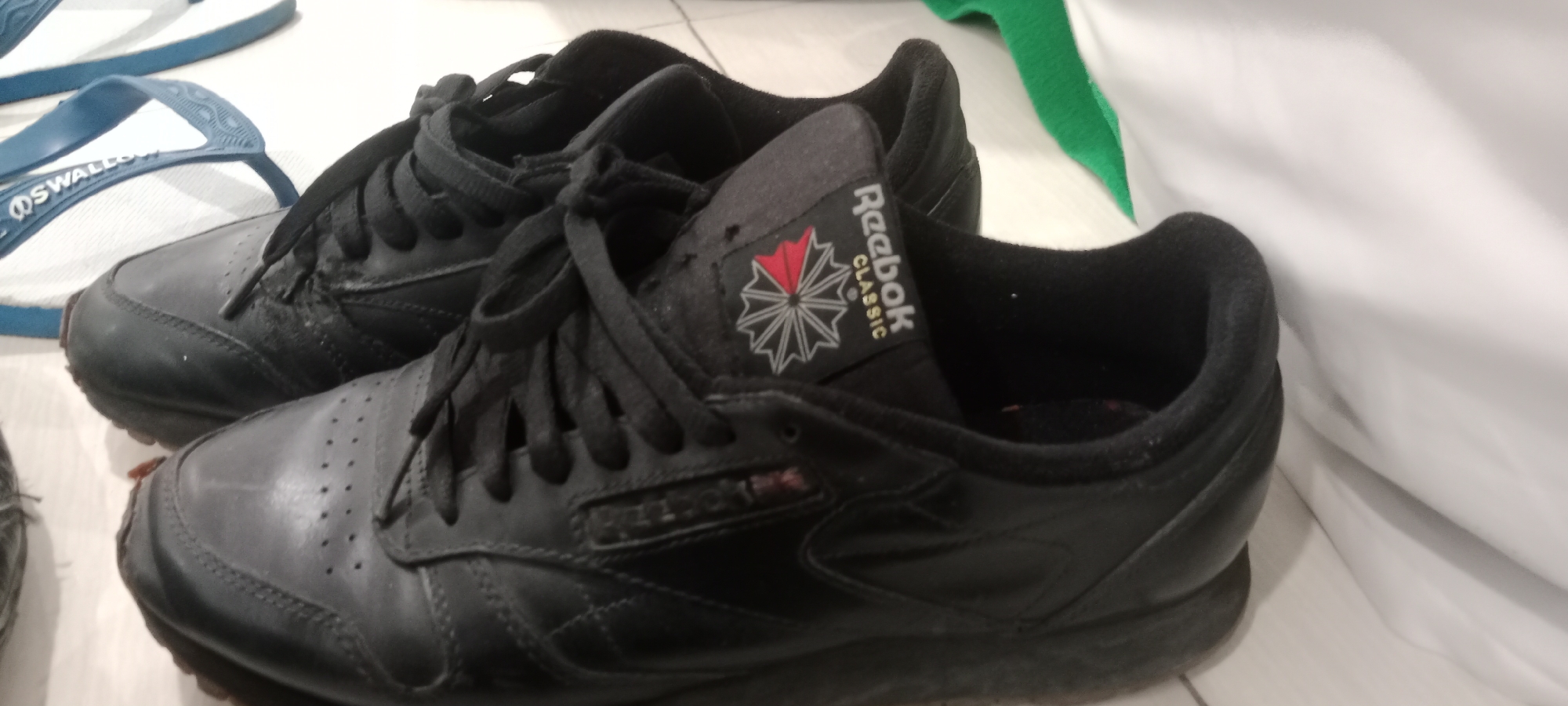 In Tuban, Indonesia, a pair of black Reebok Classic sneakers is placed on a tiled floor next to a pair of blue and white flip-flops. The sneakers are positioned in the foreground, with the left shoe slightly in front of the right. Both shoes are laced and appear to be in used condition, showing signs of wear. The tongue of the right shoe displays the distinctive Reebok Classic logo, a red and white starburst symbol next to the brand name. The flip-flops, with the brand name "SWALLOW" visible on the strap, are situated in the upper left corner of the frame, partially overlapping with the sneakers. A white fabric, possibly a garment, is draped on the right side of the image, creating a soft background. The lighting is neutral, suggesting an indoor setting.