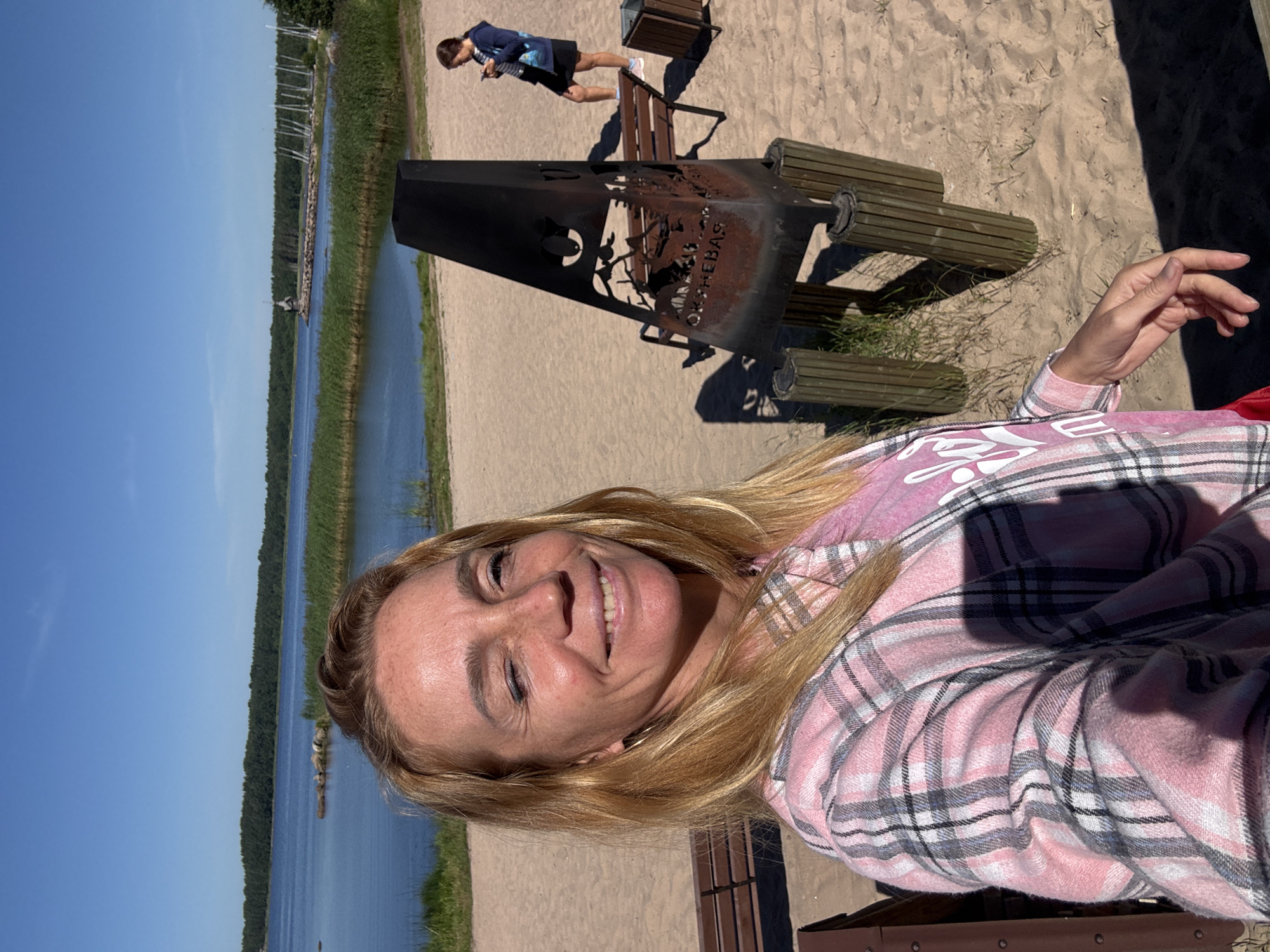 A blonde woman with freckles smiles at the camera, winking, in a selfie taken outdoors on a sandy beach. She is wearing a pink and gray plaid shirt over a pink shirt with white lettering. Behind her, a beach area with sand and sparse grass extends to a body of water. The water is calm and reflects the clear blue sky. A dense line of trees borders the far side of the water. In the middle ground, there is a black metal fire pit with a decorative cut-out design and the word "ОКНЕВАЯ" (OKHNEVAYA) visible on it. To the right of the fire pit, a woman in a dark hoodie and skirt is standing, looking at her phone. A wooden bench is situated behind the fire pit, and another bench is partially visible on the left. In the distance, a lighthouse-like structure and several sailboats are present on the water. The overall scene suggests a sunny day at a recreational waterfront location.