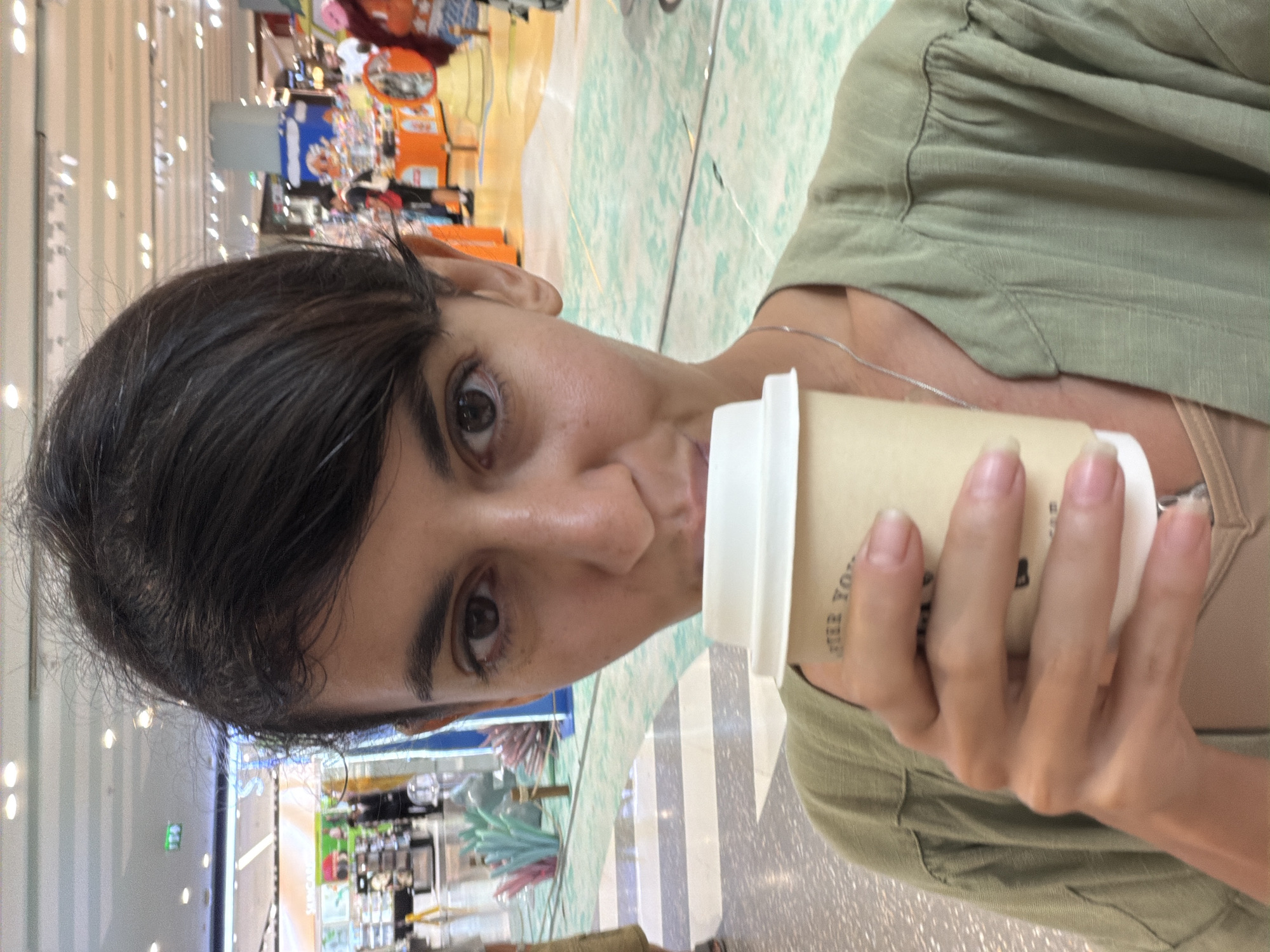 A woman with dark hair and brown eyes is holding a disposable coffee cup to her lips. She is wearing a green open-front shirt over a beige camisole. The background appears to be an indoor shopping mall with brightly colored displays and overhead lighting. The floor has a checkered pattern. The scene is likely taking place in Wichit, Thailand, a known area with shopping centers.