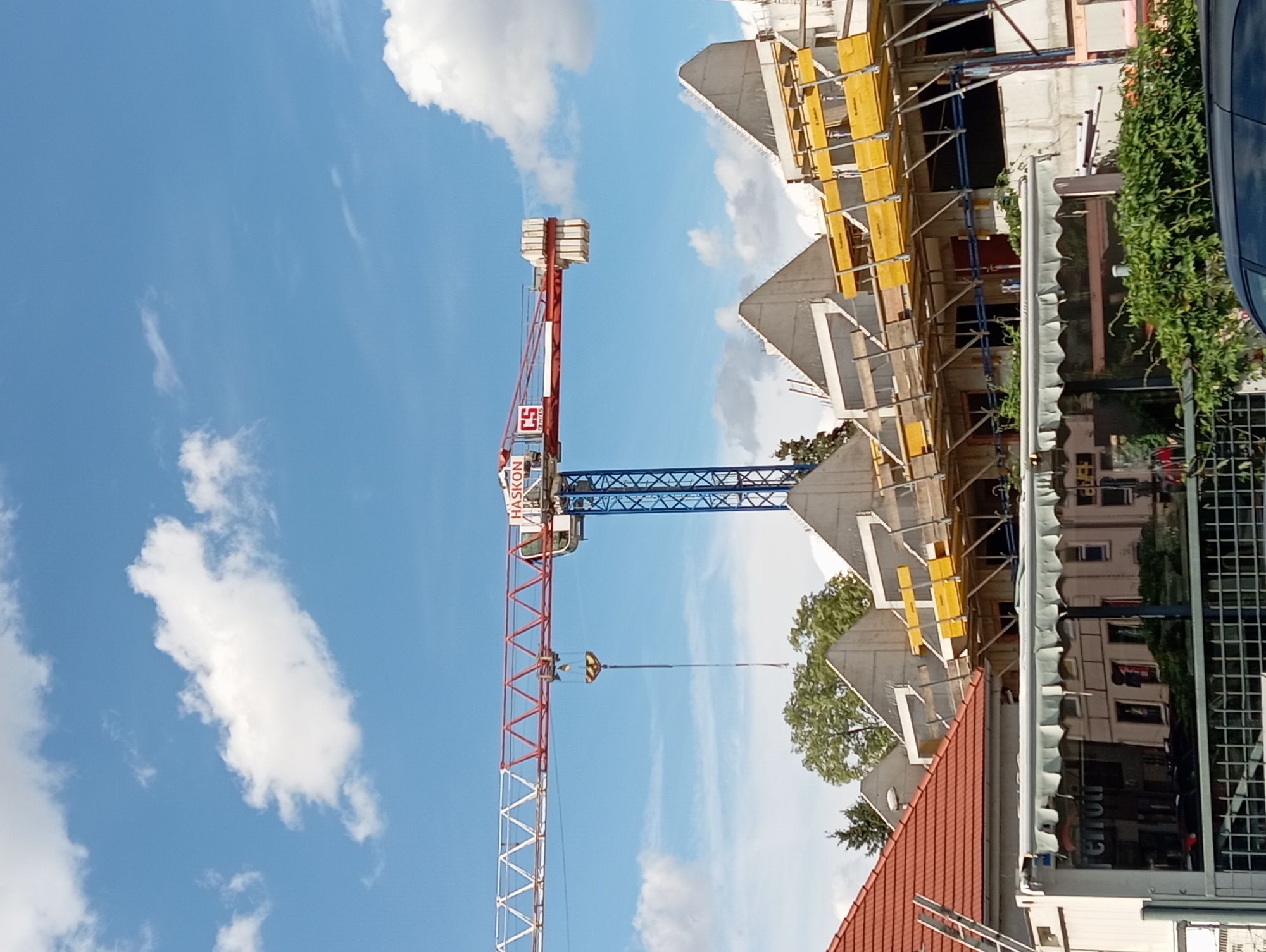 The image shows a construction site with a large tower crane dominating the frame. The crane has a red and white lattice boom and a blue horizontal structure. The cab of the crane has "HASKON" written on it, along with a red "CS" logo. The crane is positioned against a bright blue sky with scattered white clouds, suggesting it is daytime with good weather.
In the background, a building is under construction, characterized by concrete structures, scaffolding with yellow planks, and metal supports. The building appears to be multi-story with visible window openings. To the right of the construction, a dark blue car is partially visible, and some green foliage from trees is also present. The foreground shows a section of a red-tiled roof, possibly of an adjacent building. The overall scene depicts ongoing construction work in an urban or suburban setting. No people are visible in the image. The image is oriented vertically.