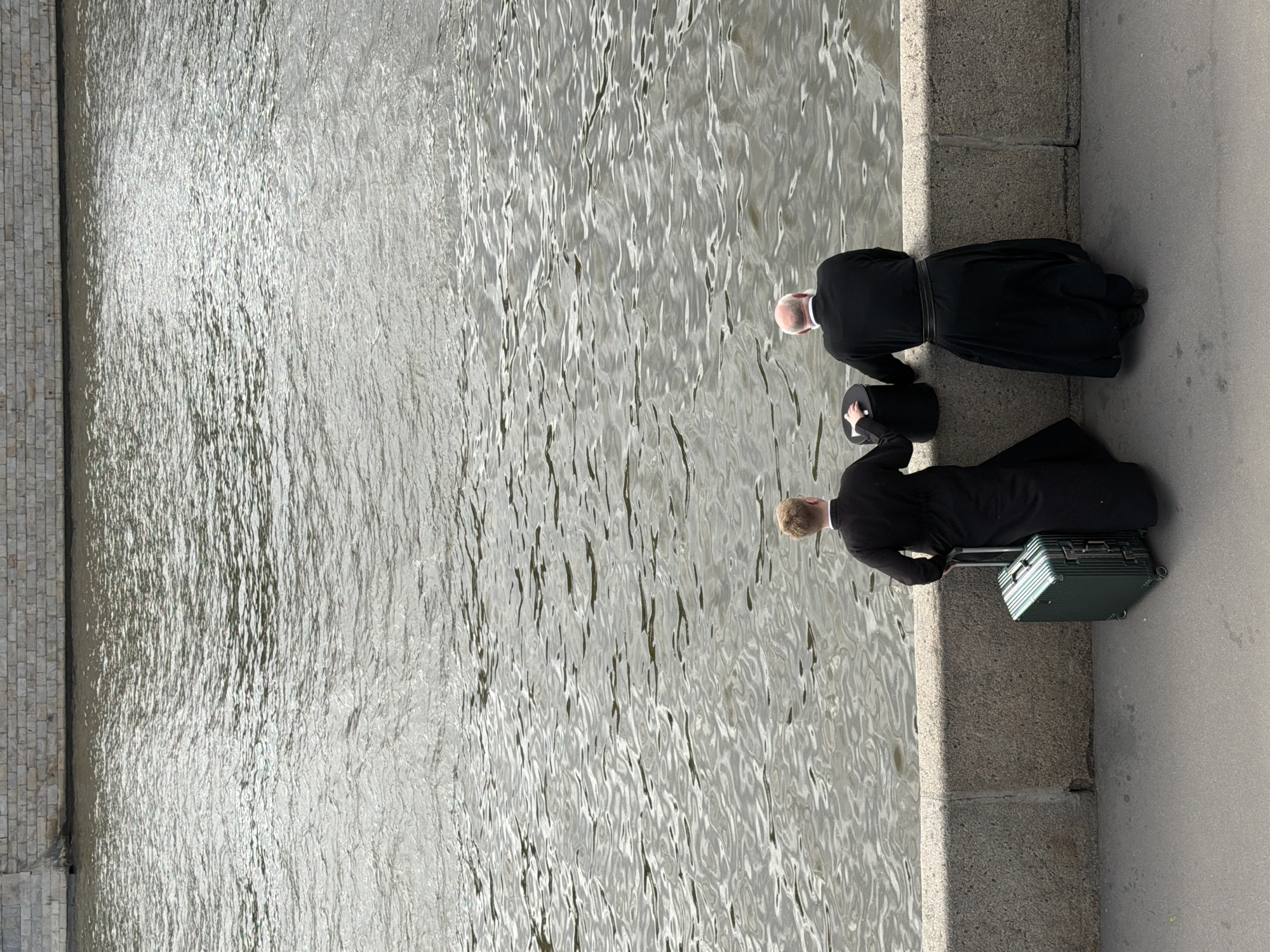 Two men in black clerical garb stand on a stone embankment next to a body of water in an urban environment. One man has a bald head and is wearing a black belt, while the other has light hair and stands next to a green suitcase. The body of water is rippled. Part of a brick wall is visible to the left. The location is Unknown, Unknown.