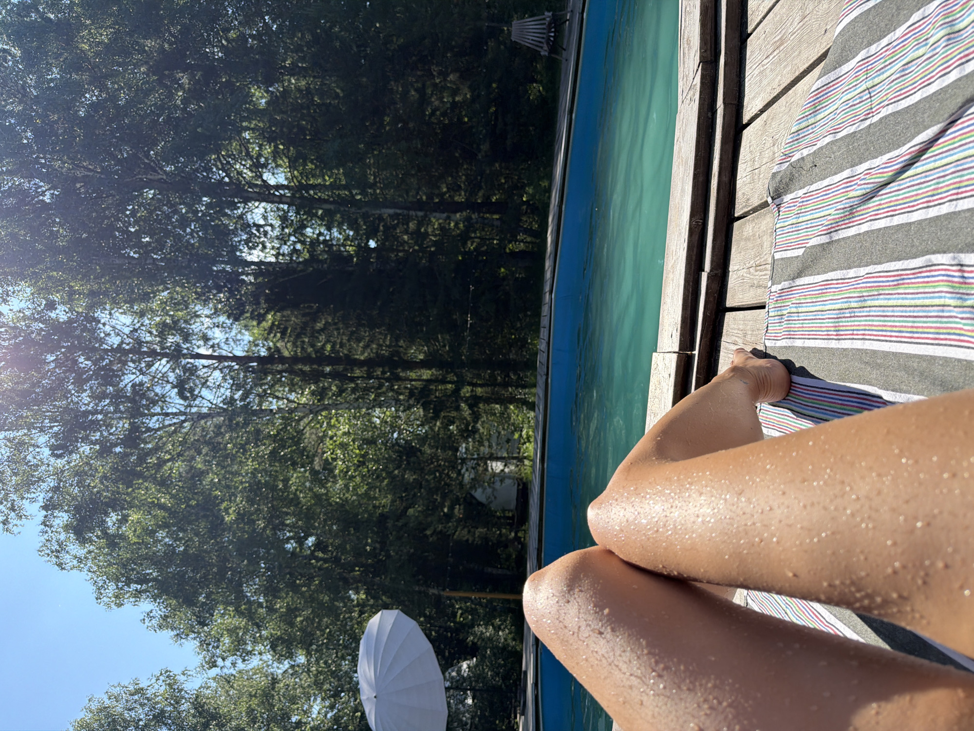 A low-angle, medium shot shows a person's legs and feet resting on a striped towel on a wooden deck next to a swimming pool. The legs are wet and glistening with water droplets. The pool water is a vibrant turquoise. In the background, a white umbrella stands to the left, and lush green trees surround the area, indicating an outdoor, possibly rural, setting. A wooden lounge chair is visible further back on the right. The sky is clear blue. This scene could be located in Miniino, Russia, given the natural, wooded environment and the presence of a pool, which are common features in suburban and rural areas of Russia during the summer.