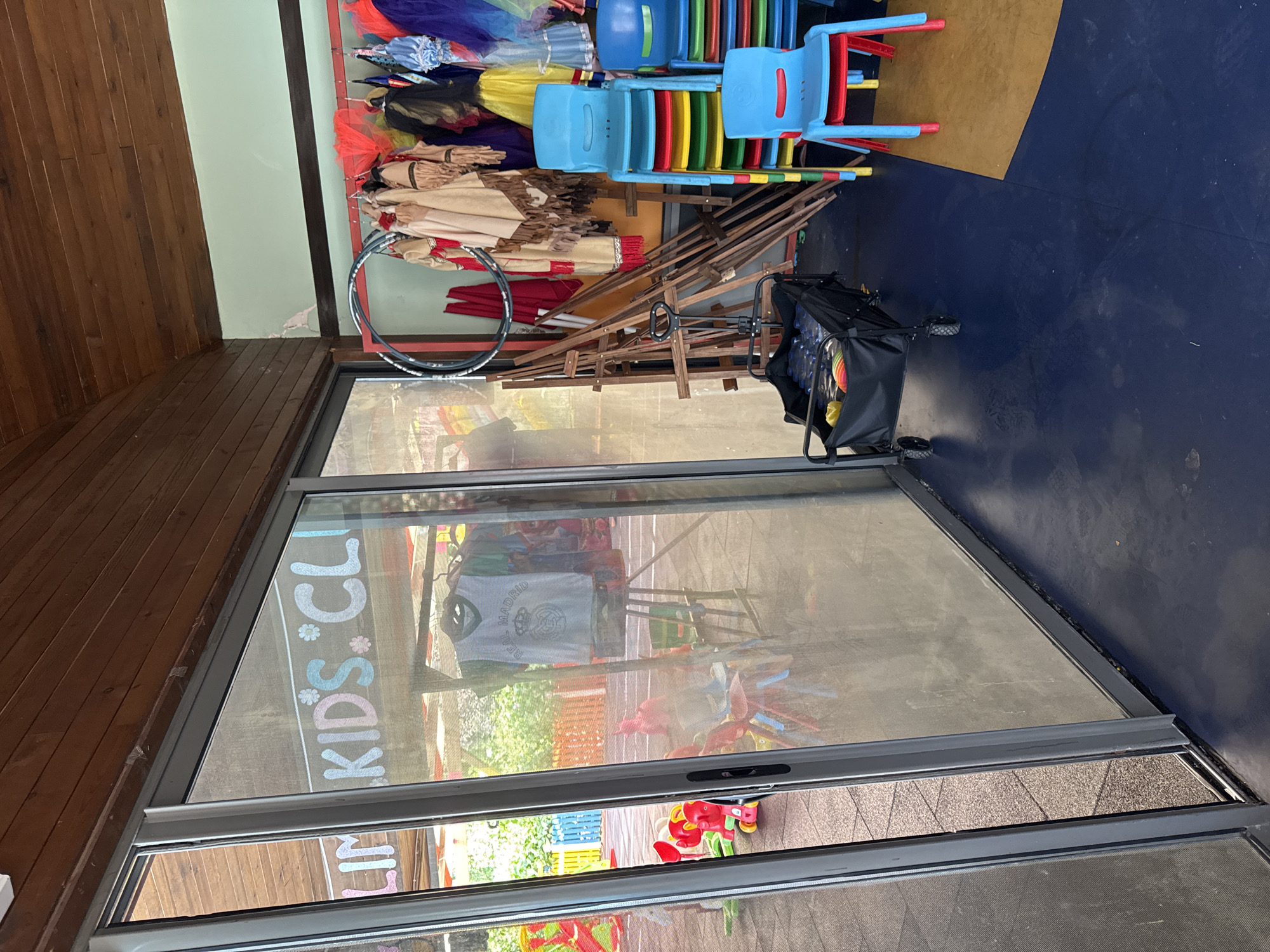 The image shows the interior of a children's club. A sliding glass door is on the left, with a mesh screen. Through the glass, a playground with colorful plastic toys and fences is visible. Inside the club, a rack of costumes hangs on the right, featuring Native American-style outfits and colorful tutus. Several stacks of bright plastic chairs are arranged near the costumes. A small black wagon filled with water bottles and a colorful ball sits on the dark blue floor. Wooden easels and a basketball hoop are also present. The walls are painted a pale green, and the ceiling is made of dark wood panels. The text "LIM KIDS' CLU" is visible through the glass door, indicating the name of the establishment. The scene is indoors and appears to be a play area or classroom for children. The location is not specified in the image or described in the prompt.