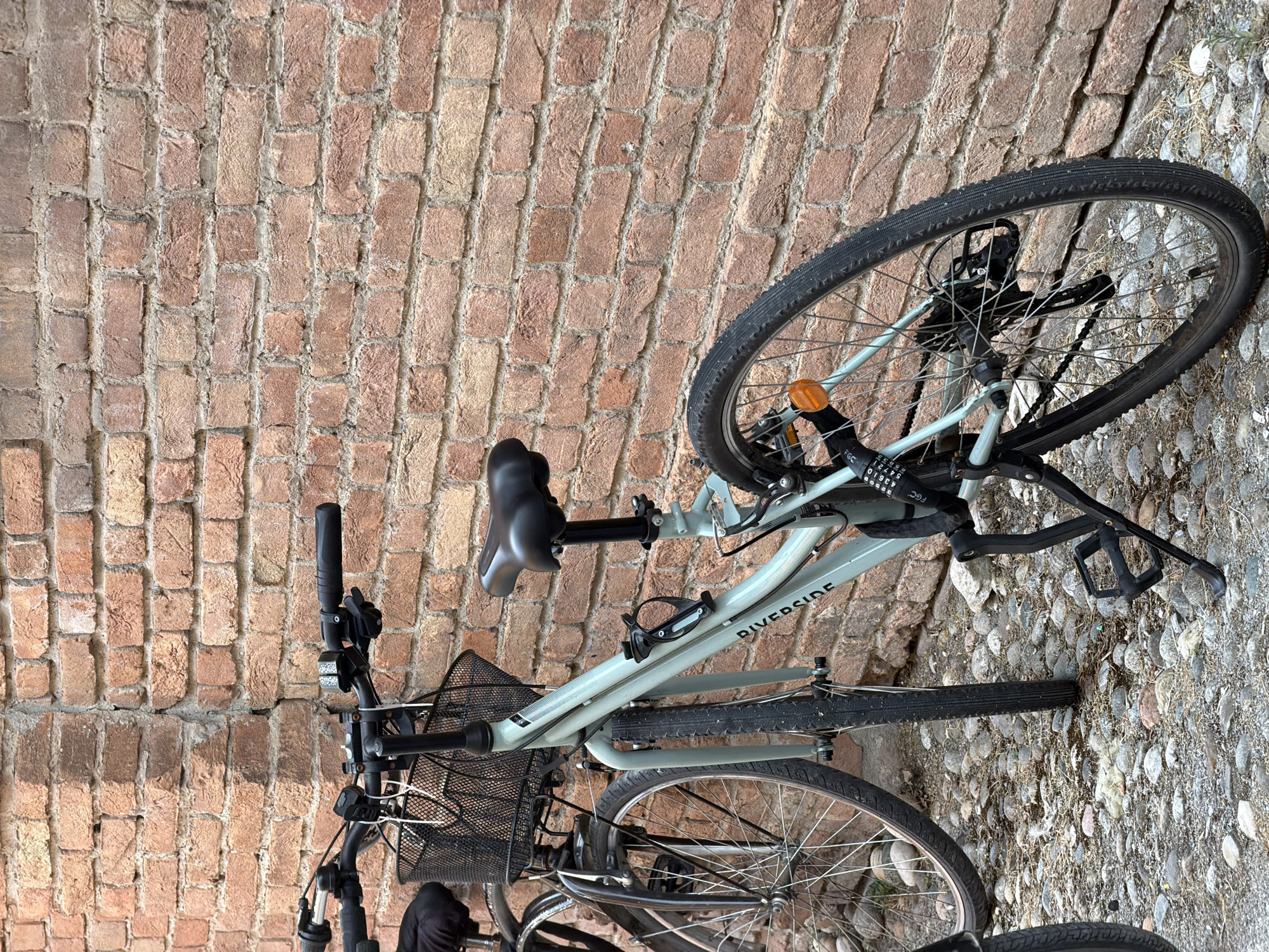Two bicycles are parked against a brick wall in Piacenza, Italy. The bicycle in the foreground is a light blue hybrid bike with a black basket on the handlebars, a black seat, and a black U-lock secured to the rear frame. The brand "RIVERSIDE" is visible on the frame. The bicycle's kickstand is extended. The ground is covered with cobblestones. The brick wall behind the bicycles is made of red bricks with visible mortar. The overall lighting suggests it is daytime. There are no people visible in the image.