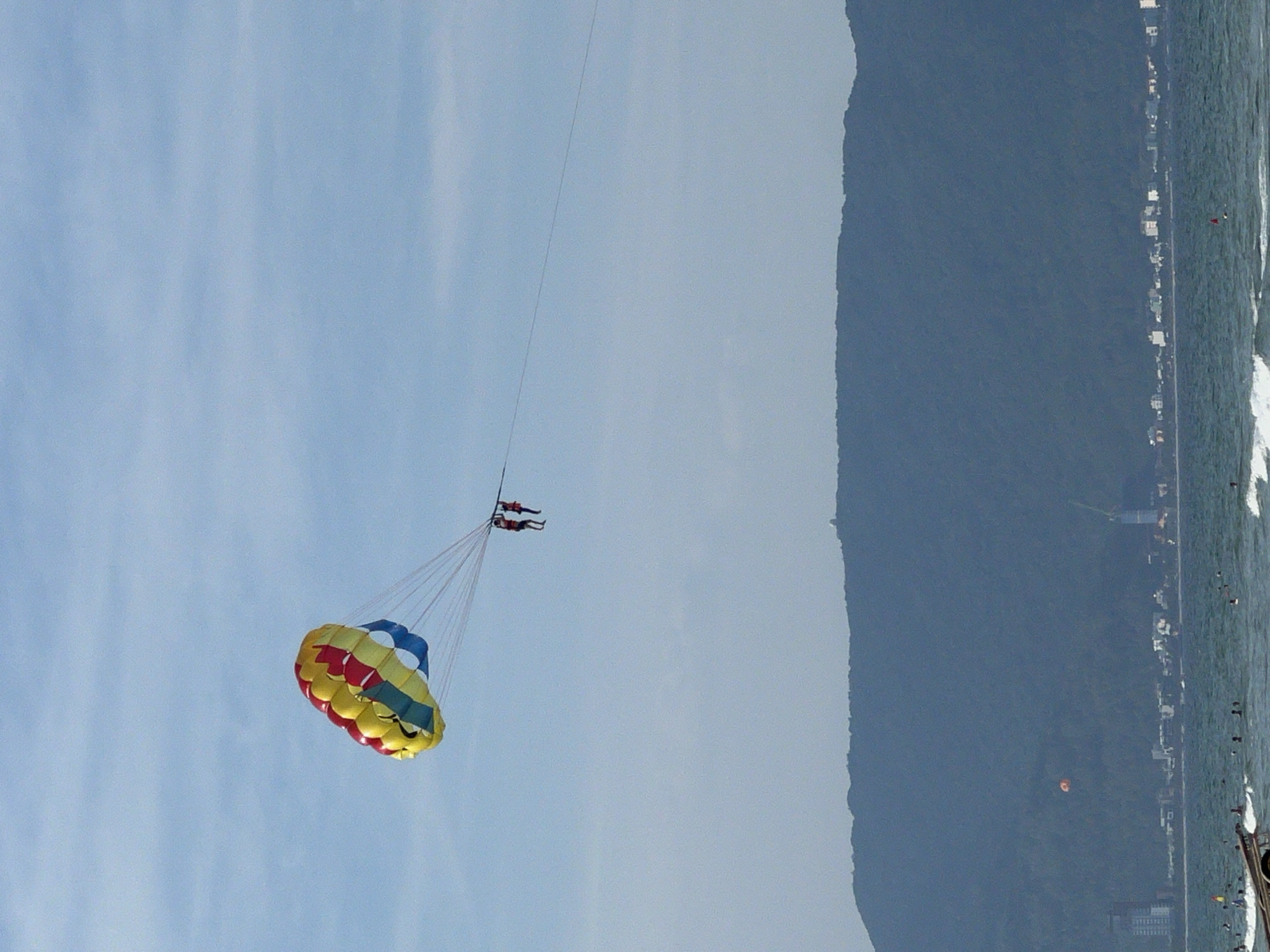 The image shows a parasailer in the sky over the ocean. The parasail is colorful, with red, yellow, and blue sections. Two people are suspended beneath the parasail. In the background, a coastline with buildings and a large mountain is visible. The ocean has waves, and there are several people in the water. The scene is likely Đà Nẵng, Vietnam, known for its beaches and coastal activities.