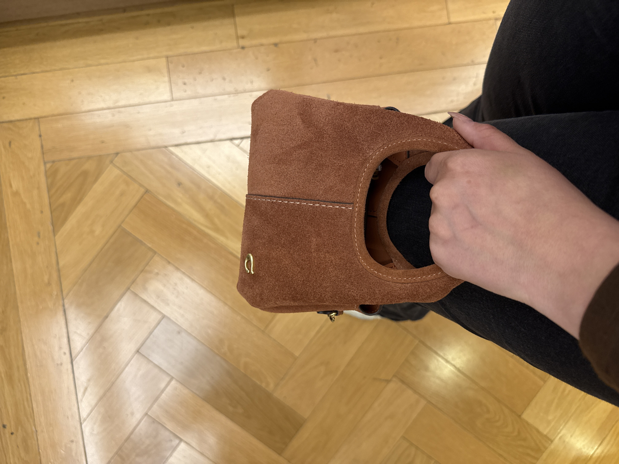 A high-angle shot shows a person sitting, holding a brown suede bag with a round handle. The person wears dark gray or black jeans. The background is a parquet wood floor, a light, natural tone. No details suggest Sunrise, United States of America.