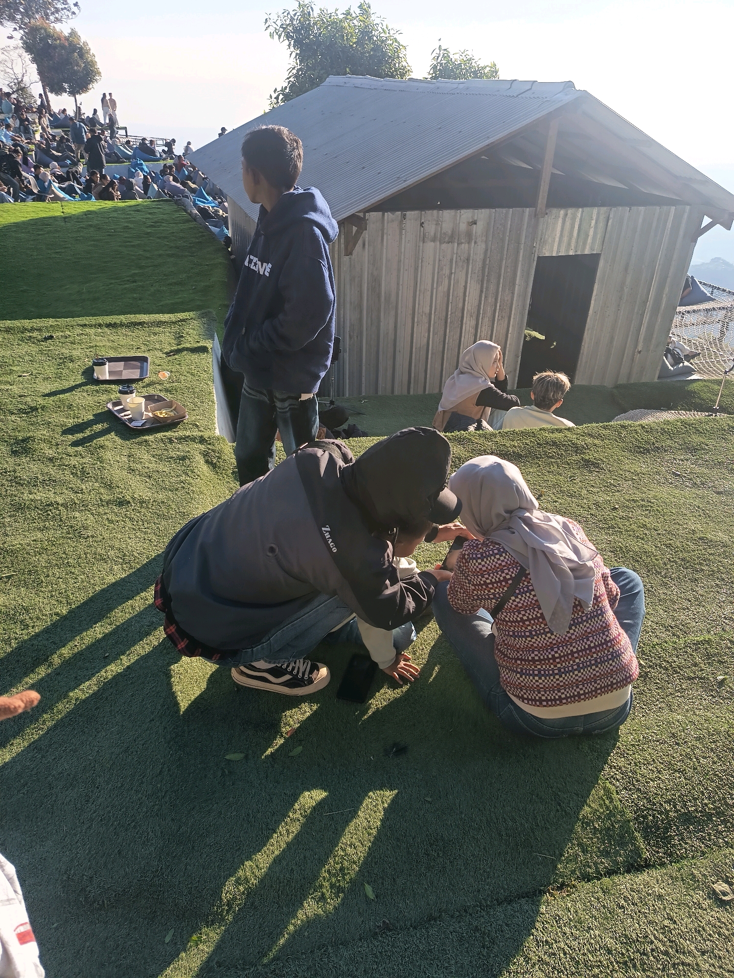 In Keditan, Indonesia, a group of people are gathered on a grassy hillside in front of a corrugated metal structure. In the foreground, two individuals are sitting on the grass, one wearing a patterned sweater and a hijab, and the other in a black hoodie and jeans, leaning forward to interact with the person beside them. Behind them, a young person in a dark blue hoodie stands and looks away from the camera. Further up the hillside, a crowd of people are seated on the grass, many in blue beanbag chairs, facing towards a distant view. The setting appears to be an outdoor seating area with a clear sky and sunlight casting long shadows across the artificial turf.