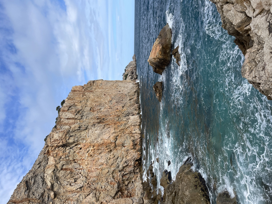 This image depicts a rugged coastal landscape in Torroella de Montgrí, Spain. A massive, vertical rock cliff, characterized by weathered tan, orange, and grey geological strata, occupies the left and central-left portions of the frame, extending from the bottom left upwards and receding into the distance. Sparse green vegetation is visible along the top edge of the cliff and on smaller landmasses further along the distant coastline. The Mediterranean Sea, exhibiting colors ranging from clear turquoise near the shore to a darker blue towards the horizon, fills the lower and right areas of the image. Waves are actively breaking against the base of the main cliff and numerous submerged and partially submerged rocky outcrops, generating white foam. Several prominent, reddish-brown and grey boulders are visible within the turbulent water. The sky above is partially cloudy, featuring a blue background with scattered white cumulus clouds. No individuals, human-made structures, or animals are present.