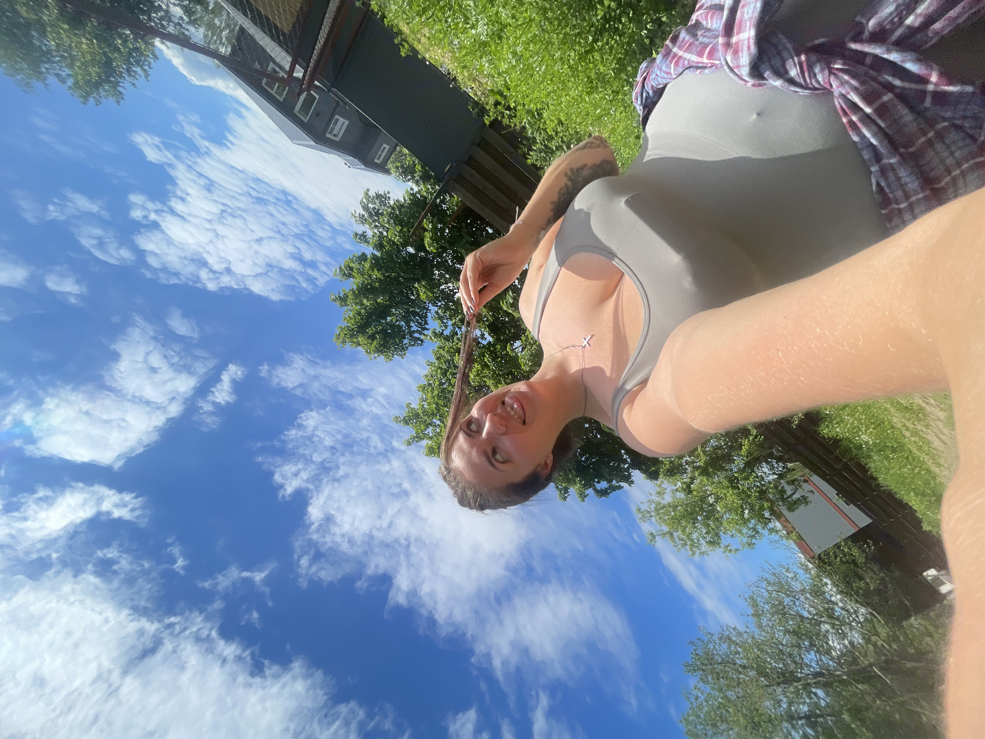 The image, originally rotated 90 degrees counter-clockwise, depicts a young woman taking an outdoor selfie on a sunny day. She is positioned on the right side of the frame, looking directly into the camera with a happy, relaxed expression, her mouth slightly open in a smile. Her long brown hair is visible, and she lightly holds a strand with her left hand, which also displays a tattoo on the forearm.
She is wearing a light grey tank top or sports bra, revealing a delicate necklace with an 'X' pendant. A red and blue plaid shirt is tied around her waist. The background showcases a vibrant outdoor environment with a clear blue sky dotted with fluffy white clouds, indicating bright daylight. Below the horizon, green grass and several leafy trees are visible. A dark-colored house or building with white-framed windows can be seen in the upper right background, partially obscured by foliage. No text is visible in the image. The scene conveys a casual and pleasant outdoor moment.