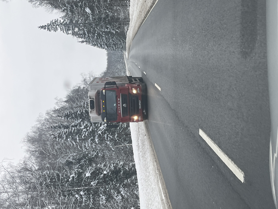The image captures a winter road scene, specified as being in Butyrki, Russia. An overcast sky casts a muted light over the environment. A red SITRAK brand tractor-trailer truck, featuring a white refrigerated unit on its trailer, is prominently positioned in the center, traveling towards the viewer. Its headlights and fog lights are active, and a plume of exhaust is visible above the cab. A Russian license plate is affixed to the truck's front bumper. The two-lane asphalt road appears dark and damp, marked with white dashed lines separating the lanes and a solid white line defining the right shoulder. Substantial snow banks line both edges of the road. The surrounding landscape consists of a dense forest with snow-laden evergreen trees and bare deciduous trees, extending into the background where the road gently curves to the right. The perspective suggests the photograph was taken from inside another vehicle, with a portion of its interior visible at the bottom edge of the frame.