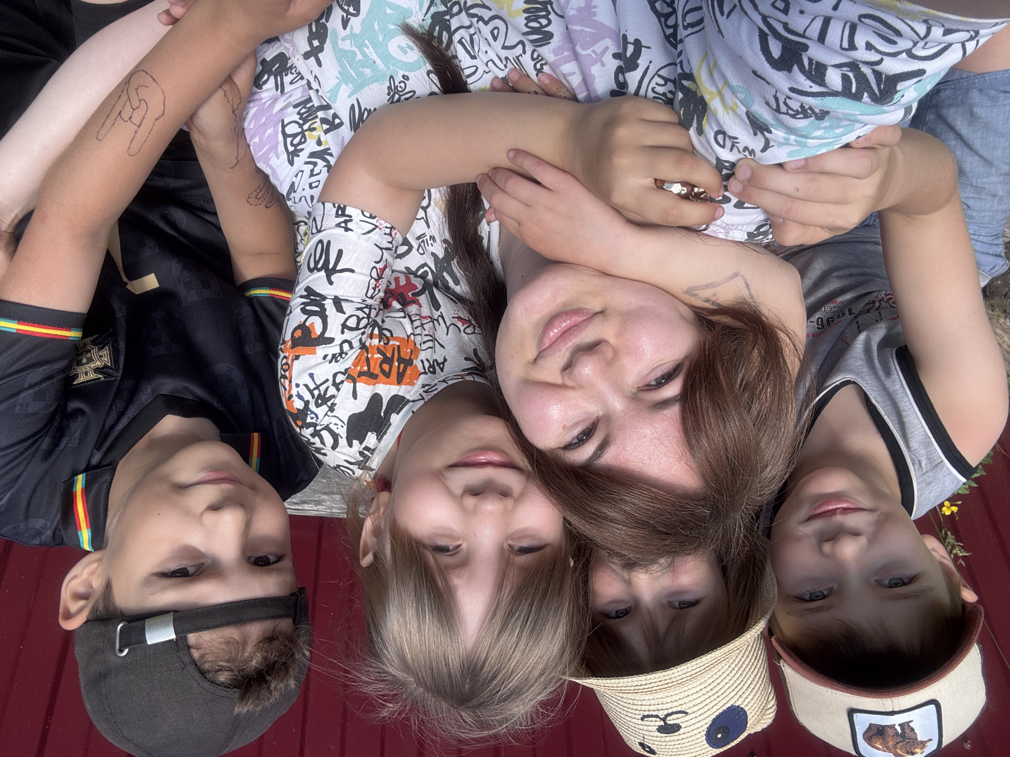 A group of five people, four children and one adult woman, are posing for a selfie outdoors. The woman is in the foreground, with her arms around two of the children. The children are of varying ages, and they are all smiling. One of the children is wearing a baseball cap with a bear graphic, another is wearing a straw hat with a panda design, and the third is wearing a black baseball cap. The adult woman has a tattoo on her arm. The background is a red wooden wall. The scene appears to be in a residential area, possibly a backyard.