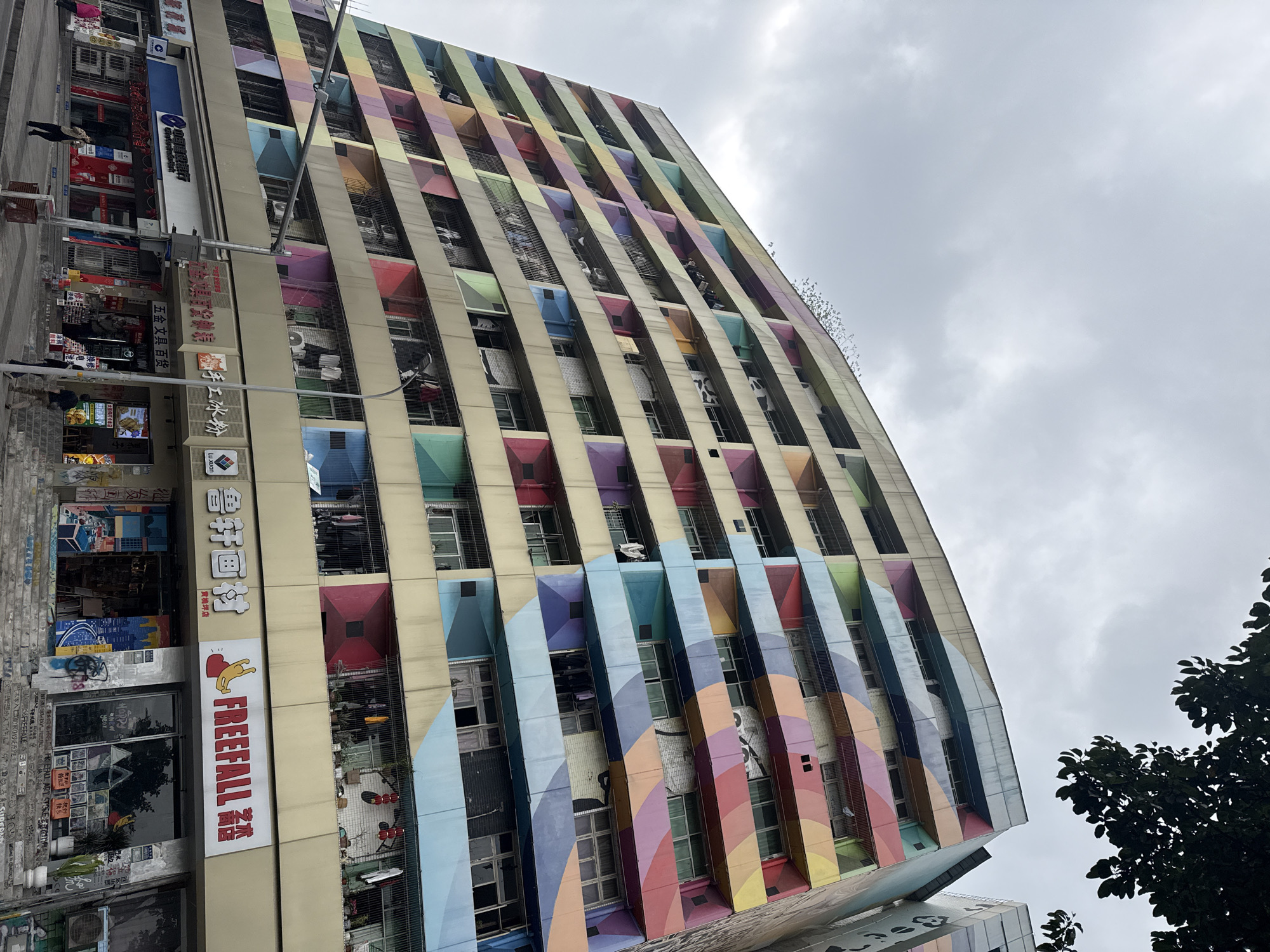 The image displays a multi-story residential and commercial building situated in Jiulongpo District, China, under an overcast sky. The building's facade is distinctive, featuring a vibrant, multi-colored geometric pattern composed of horizontal and diagonal bands in shades of blue, green, yellow, orange, red, and purple, applied over a beige base. Numerous windows and enclosed balconies are visible across at least ten floors. Clothes are observed drying on several balconies, and some windows are open. A flock of approximately two dozen small birds is flying near the upper left corner of the building, above the rooftop. A tree branch with green foliage extends into the upper right corner of the frame.
At street level, the building houses various commercial establishments. Visible storefronts include a bank with a prominent blue circular logo, several shops with Chinese signage such as "手工冰粉" (Handmade Ice Jelly), "五金文具百货" (Hardware Stationery Department Store), and a "鲁轩画材" (Lu Xuan Art Supplies) store. Adjacent to the art supplies store is a "FREEFALL" art store, identifiable by its English name, a yellow figure logo, and graffiti around its entrance. Multiple individuals are present on the sidewalk: one person in dark clothing walks left near the bank, another stands near the steps of a shop to the right, and other pedestrians are partially visible. A street light pole with attached signal equipment is positioned on the right side of the frame, obscuring a portion of the building's lower floors.