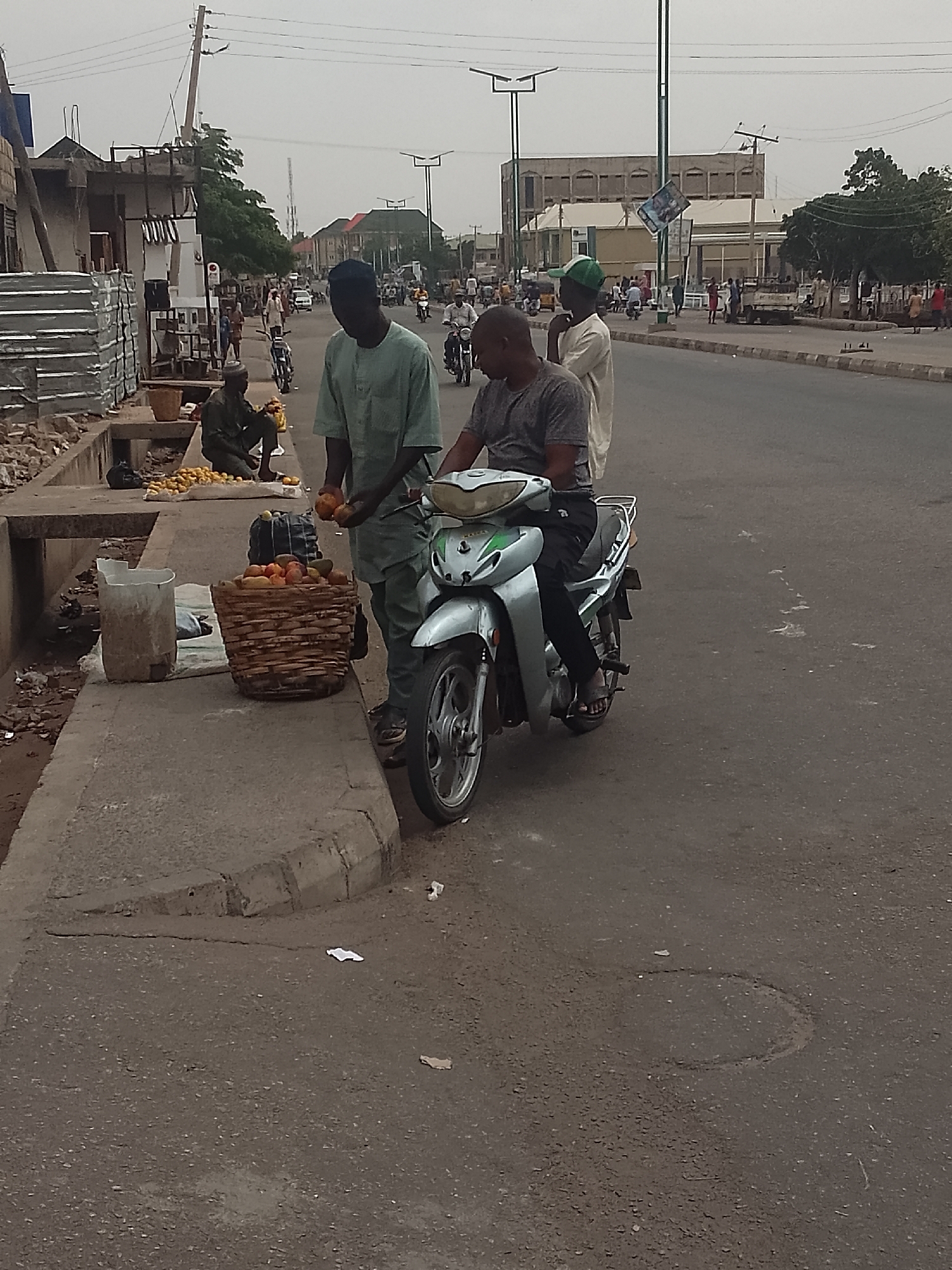 The scene depicts a street in Pantame, Nigeria. A man in a light green tunic is standing on the sidewalk while interacting with another man sitting on a silver motorbike parked on the road. The man on the bike is wearing a grey shirt and black pants. On the sidewalk, next to the man in green, is a woven basket filled with reddish-orange fruits. Near the basket, a white bucket sits on the sidewalk. Further down the sidewalk, another man is sitting on a ledge with piles of yellow fruits spread out around him. The area appears to be a commercial zone with buildings visible in the background and pedestrian traffic on the sidewalk. Other motorbikes and vehicles are also on the road. Streetlights line the street. The sky is overcast.