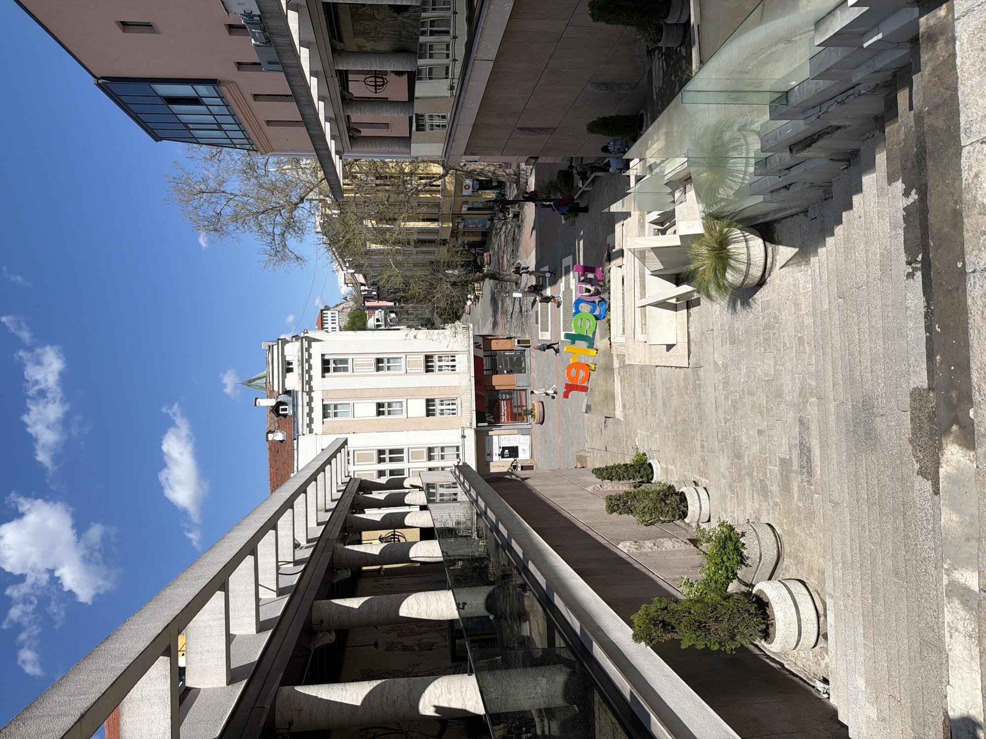 The image presents an elevated view of a bustling urban pedestrian street, likely in Plovdiv, Bulgaria. The scene is dominated by a clear blue sky with sparse white clouds, indicating sunny weather during the day, possibly mid-morning or afternoon, given the distinct shadows.
The street is lined with multi-story buildings, some featuring light-colored facades and large windows, while others in the background appear older with pitched, tiled roofs. Several bare deciduous trees are interspersed along the street. On the right side of the frame, a modern elevated platform or terrace is visible, equipped with a concrete balustrade and supported by concrete columns. This elevated area is separated from the street by a transparent glass barrier, and a broad set of stone steps descends from it to the street level. Planters with green, bushy vegetation are placed along this elevated section and near the steps.
At street level, several people are casually walking or gathered. A prominent feature is a large, colorful, three-dimensional sculpture spelling out the word "Together" on the paved surface of the street. The ground is primarily paved with stone or concrete slabs. The overall impression is one of a vibrant and active public space on a pleasant day. No other specific legible text is visible apart from the large letters "Together".