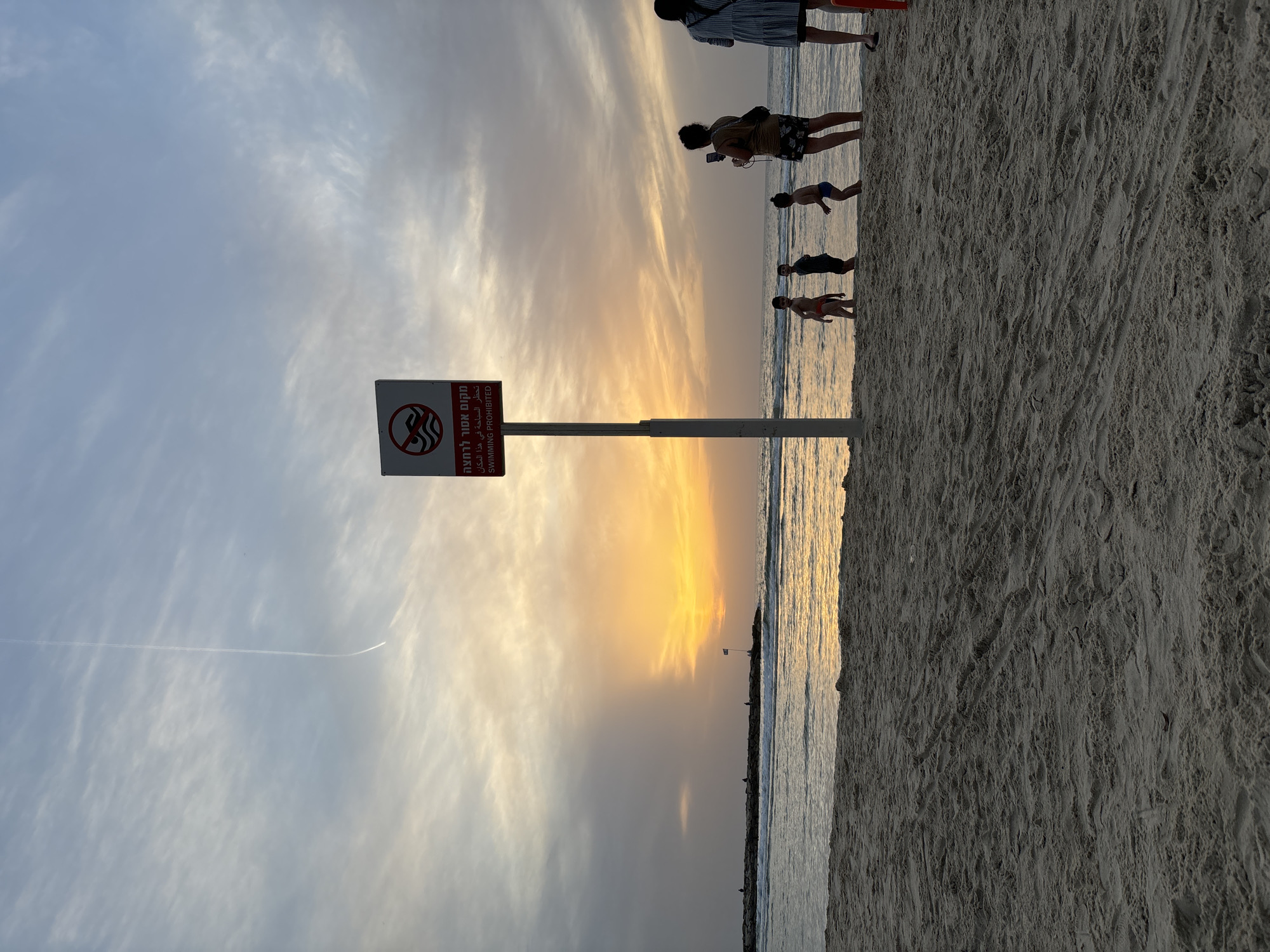 The image depicts a beach scene at sunset, likely in Tel Aviv, Israel. The sky is a mix of light blue and orange hues. A no-swimming sign stands prominently in the foreground. Several people are seen at the water's edge. The beach is sandy, and there are waves in the sea. A contrail is visible in the sky.