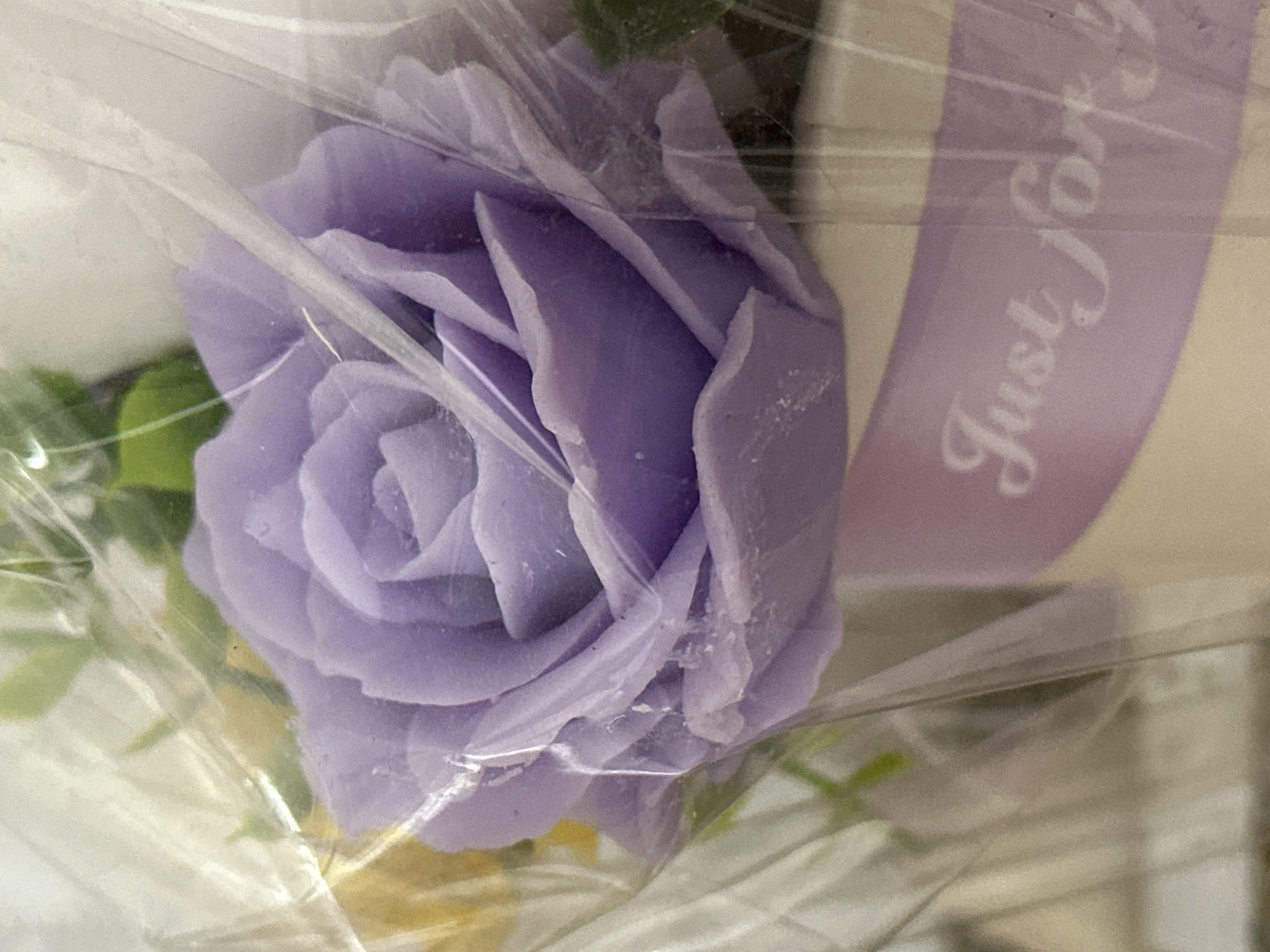 The image shows a close-up view of a purple rose, possibly made of soap or paper, enclosed in clear plastic. The rose is in full bloom with many petals. Below the rose, a light purple ribbon with the white cursive text "Just For You" is visible, wrapped around what appears to be a container or vase. The background is blurred and out of focus, with hints of green foliage and other flowers. The lighting is soft, and there are some reflections on the plastic wrapping. The image does not contain any discernible information that would allow for its location in Yessentuki, Russia, or any specific environmental context beyond the immediate floral arrangement. There are no people or actions depicted in the image.