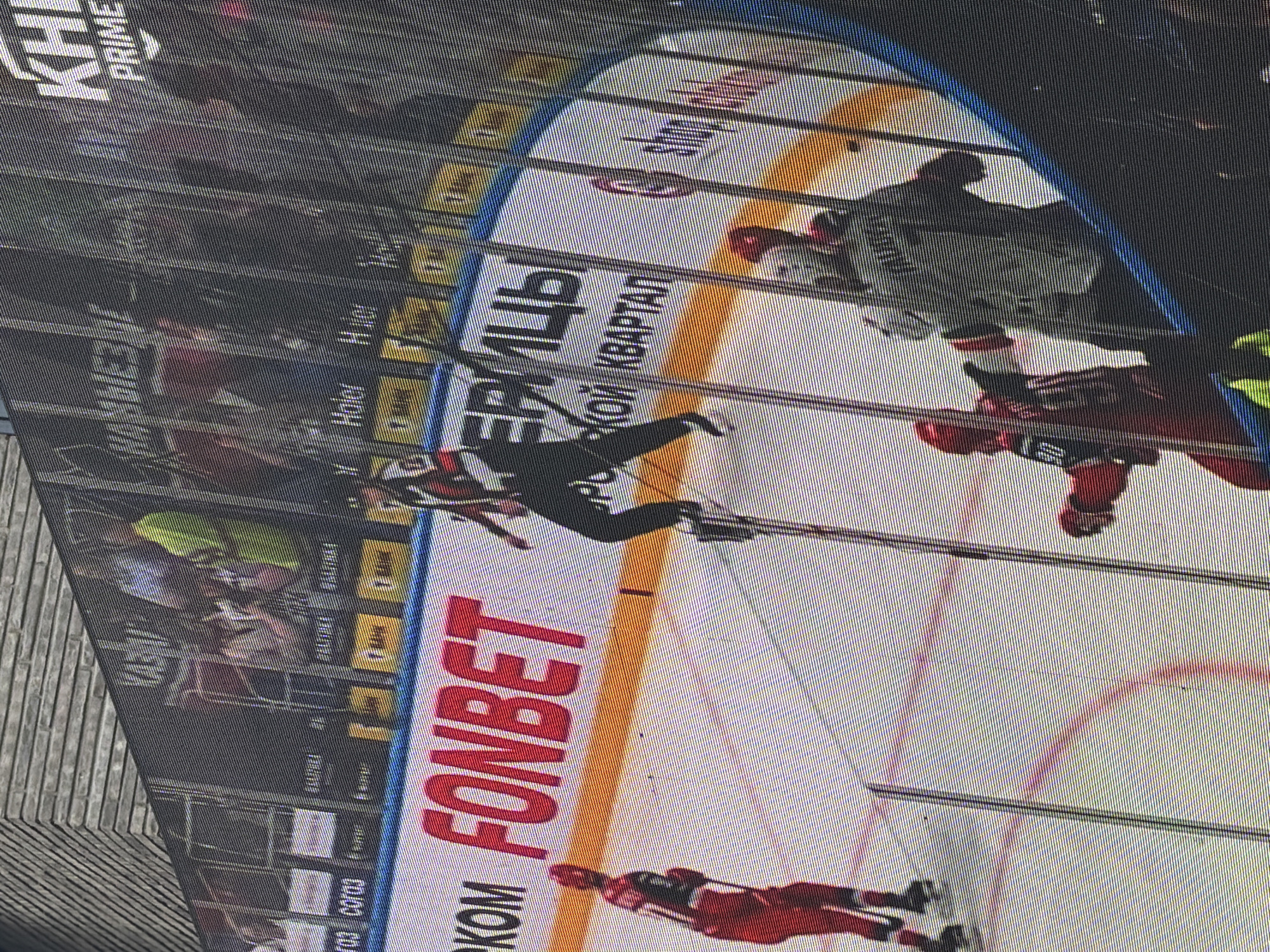 The image shows an ice hockey game in progress, captured through a barrier, possibly a glass panel or a screen. The arena features advertisements, including "FONBET" in large red letters and "ЕРМИЦЫ, ЯКОЙ КВАРТАЛ" above it. Multiple hockey players, dressed in black and red uniforms, are visible on the ice, engaged in the game. The background includes spectators and additional signage, including "KH" and "PRIME." Given the signage and the city mentioned, it's likely a game of Ak Bars Kazan, a professional ice hockey team based in Kazan, Russia. The specific location would be the arena where the game is taking place.
