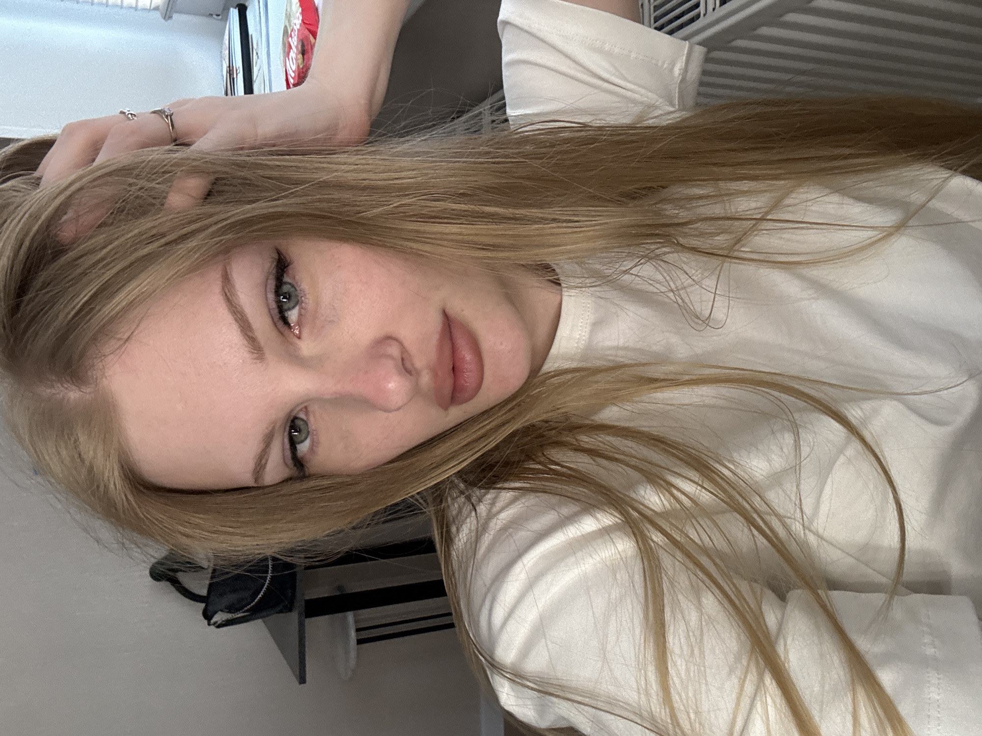 A young woman with long, blonde hair is lying down in what appears to be a bedroom. She is wearing a white t-shirt. Her hair is spread out around her face and over her shoulders. She is wearing makeup, including eyeliner and mascara, and has two rings on her left hand. The lighting suggests it is daytime. There is a shelf with some items on it visible in the background, and a radiator. The overall mood is casual and relaxed.