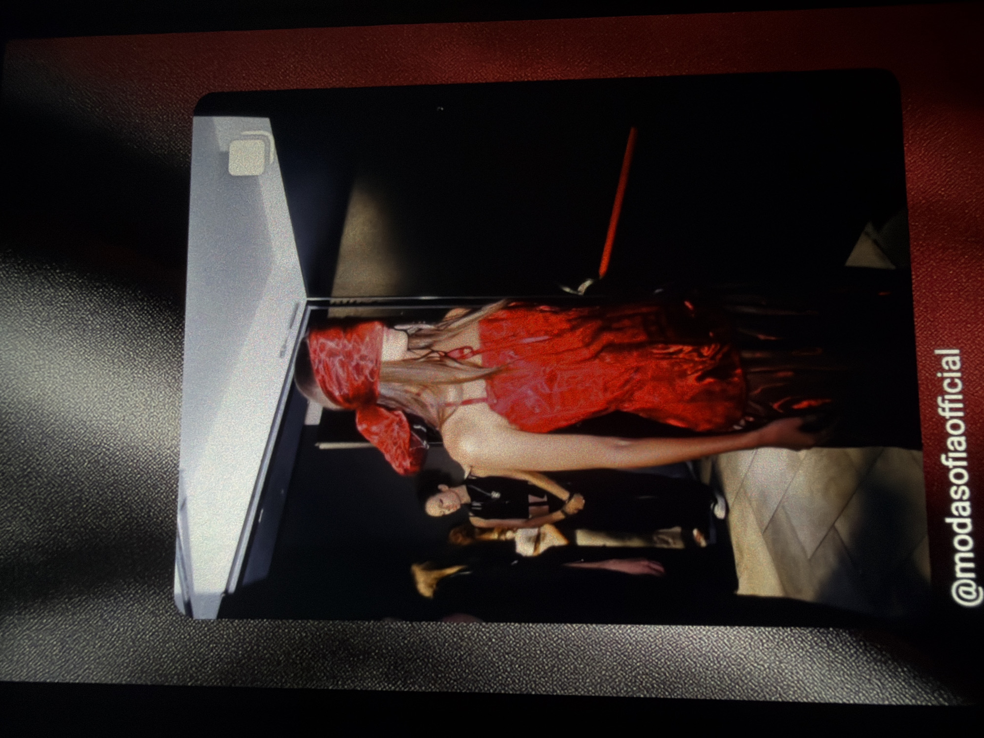 A person wearing a red corset and a red blindfold stands in what appears to be a backstage area of a fashion show. They are illuminated by stage lighting, with a dark backdrop and other individuals in the background, some of whom are also dressed in dark clothing. The floor is tiled. A red bar or rod is visible horizontally in the mid-ground. A social media handle "@modasofiaofficial" is overlaid at the bottom of the image. The image is a photograph of a screen displaying the scene, with a textured border. The location in the city of Sofia, Bulgaria is not discernible from the visual information provided.