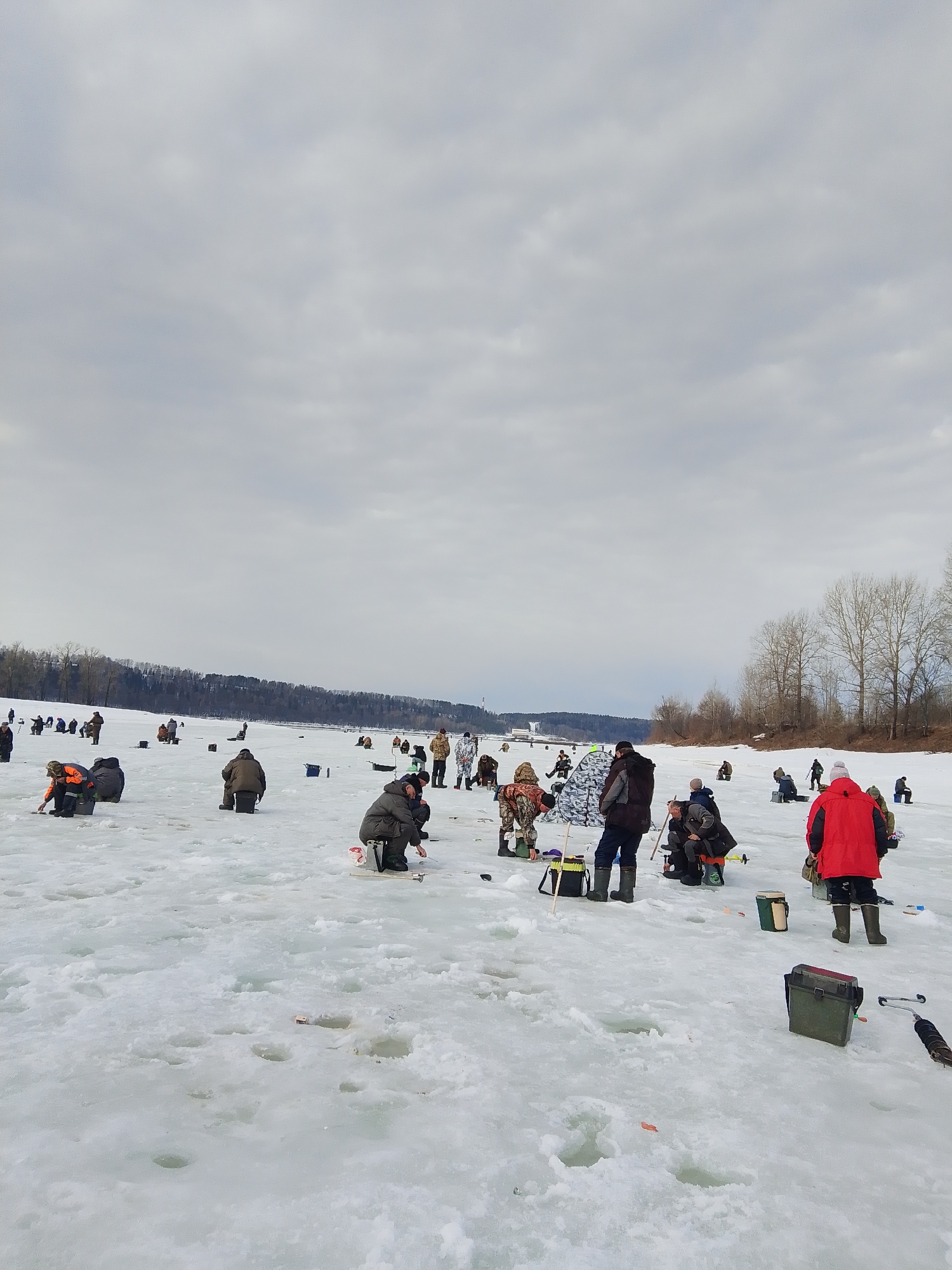 The image shows a group of people ice fishing on a frozen body of water. The location is Ol'khovskoye, Russia. The ice is covered in snow and has numerous holes drilled into it. People are scattered across the ice, some sitting on stools or kneeling by the holes, seemingly engaged in fishing. They are dressed in winter clothing, with various jackets, hats, and boots visible. Several fishing tools and equipment, such as ice fishing rods, tackle boxes, and bags, are scattered around. The background features a line of trees along the shoreline under an overcast sky.