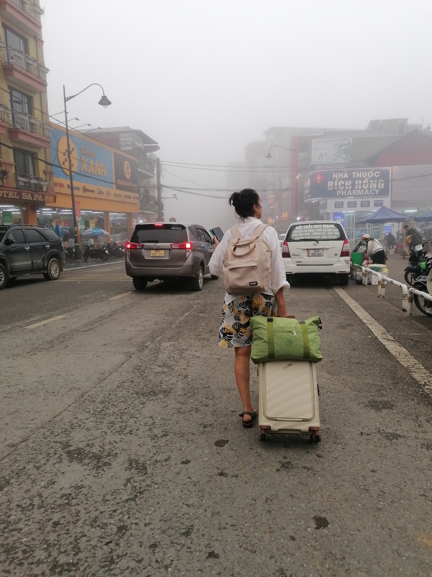 A rear view captures a person walking along a fog-laden street in Sa Pa Ward, Vietnam, carrying luggage. The central subject is a person, likely female, with dark hair tied back, wearing a white long-sleeved shirt, patterned shorts, and sandals. They are carrying a beige backpack with "SUNNY" written on it and pulling a light-colored rolling suitcase with a green duffel bag placed on top.
The street appears to be an urban thoroughfare, with buildings lining both sides. Heavy fog permeates the atmosphere, significantly reducing visibility and giving the scene a muted, somewhat dreamlike quality. Vehicles, including a dark SUV, a brown sedan (license plate 29H 957.33), and a white sedan (license plate 30M 382.84), are present on the road. Several motorbikes are parked or moving in the background.
Visible signage on the buildings indicates commercial activity. On the left, a large sign reads "Dienmay XANH" and "thegioididong," identifying an electronics store. Partial text "OTEL SA PA" suggests a hotel. On the right, a prominent blue sign displays "NHÀ THUỐC BÍCH HỒNG PHARMACY," indicating a pharmacy. Another sign further down reads "NỘI THẤT DUY MẠNH" with contact numbers, advertising a furniture business specializing in interior design. Street lamps and overhead wires are also visible.
The activity suggests travel or commuting through the foggy town, possibly arriving at or departing from a hotel or residence. The time of day is difficult to ascertain due to the dense fog, but it appears to be daytime. The overall impression is an everyday scene in a Vietnamese town, characteristic of the misty conditions often experienced in Sa Pa.