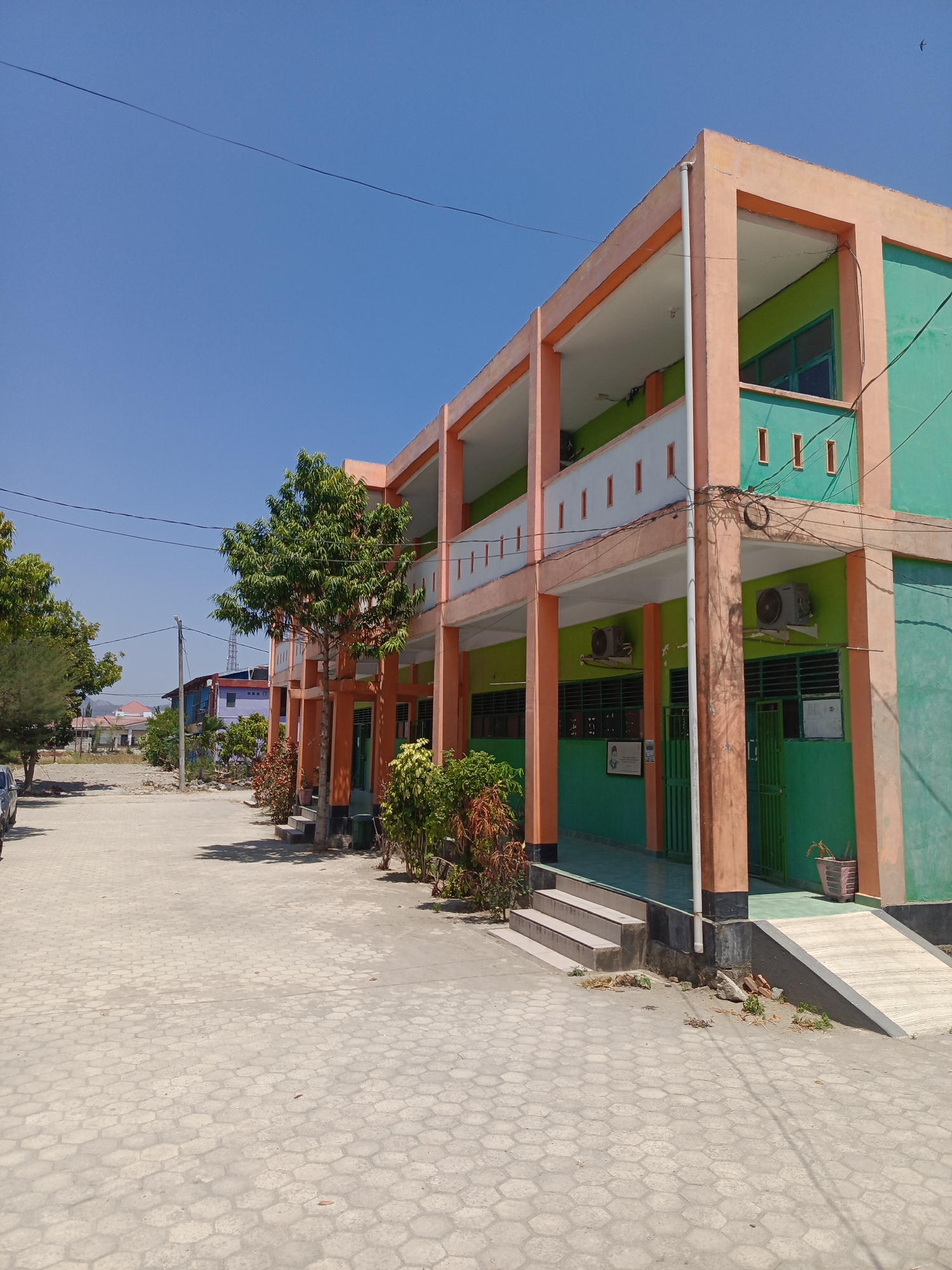 The image depicts a school building with a distinct coral-orange and emerald-green color scheme, situated in Majelling Watang, Indonesia. The building has two stories, with balconies on both levels supported by coral-orange pillars. The ground floor features multiple windows with dark green bars and doors, also in dark green. Air conditioning units are visible mounted on the walls of the second floor. The foreground is a paved walkway with hexagonal concrete tiles. To the left of the building, a row of trees stands, and beyond that, a dirt area leads to other structures, including a blue and purple building. The sky is clear and blue. There are no people or animals visible in the image. The scene appears to be an outdoor educational facility on a bright, sunny day.