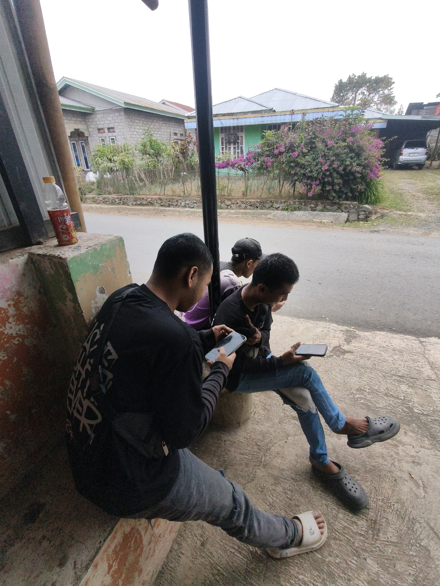 Three young men are sitting on a concrete bench outdoors. The man in the foreground is wearing a black long-sleeved shirt and ripped blue jeans, with a white sandal on his right foot and a black clog on his left. He is looking down at a smartphone in his hands. The man to his right is wearing a black t-shirt, blue jeans with a ripped knee, and black clogs. He is also looking at a smartphone. The third man, in the background, is wearing a black cap and a purple shirt, and appears to be looking at his phone as well. A bottle of Pucuk Harum tea is on a concrete surface to the left of the man in the foreground. In the background, there are houses with metal roofs, some with purple flowering bushes in front of them. A silver car is parked under a carport on the right side of the image. The ground is a paved road and a concrete walkway. The location is identified as Lindung Jaya, Indonesia.
