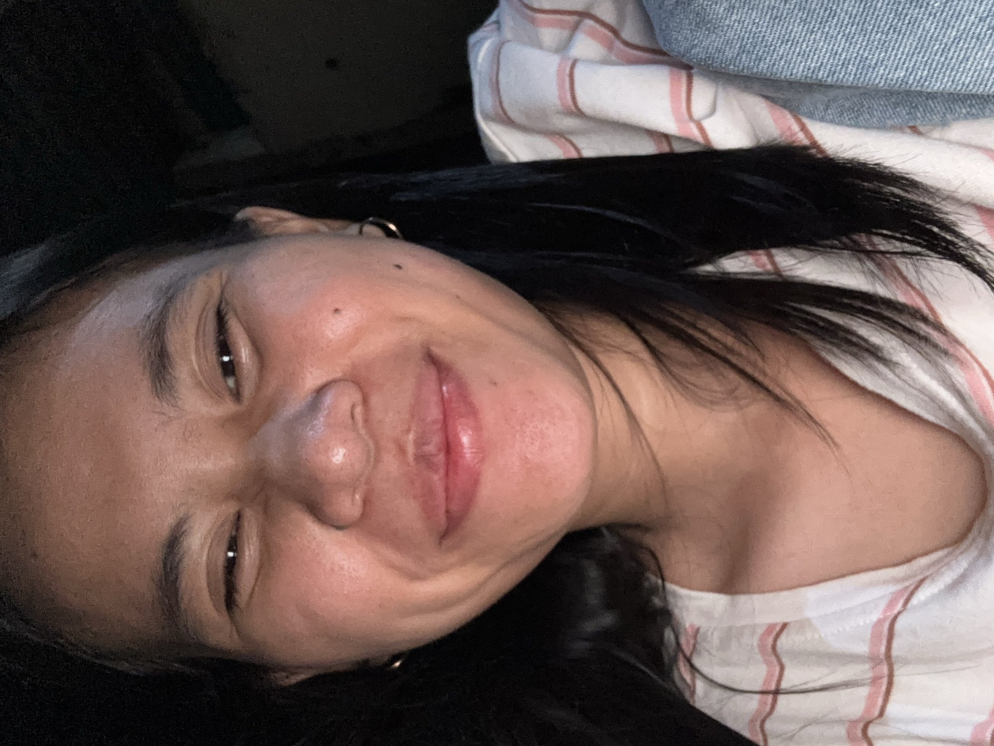 The image shows a close-up, selfie-style shot of a young woman with dark hair and light brown skin. She is smiling with her eyes squinted and her lips parted. She is wearing a white t-shirt with pink stripes. She has small hoop earrings in both ears and a beauty mark on her right cheek. The background is dark and out of focus, suggesting an indoor setting with low lighting, possibly a car interior given the angle and the hint of a denim jacket on the right side of the frame. There is no identifiable information about her location within East Jakarta, Indonesia.