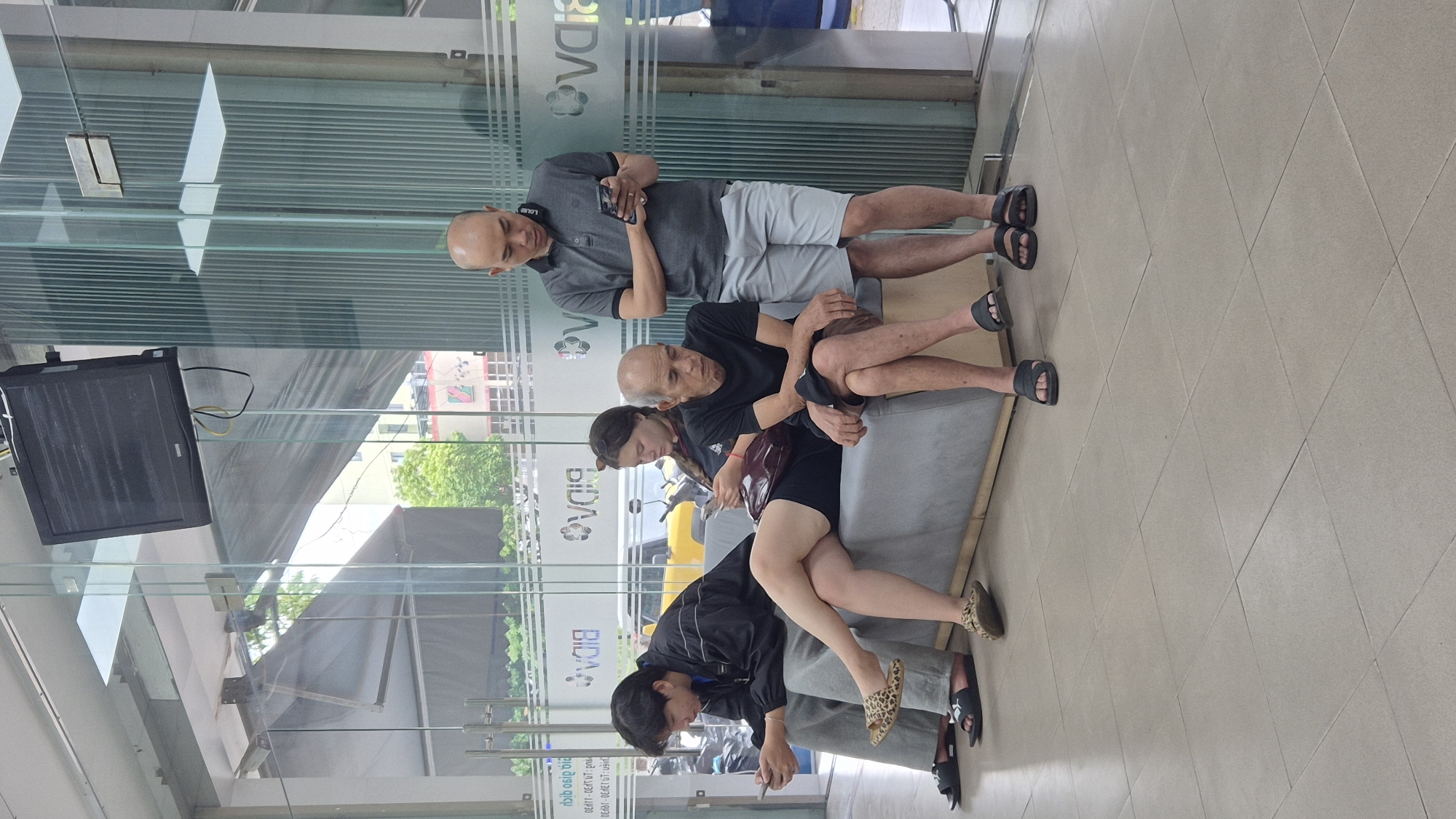This image shows four individuals waiting in a brightly lit, modern interior, likely a bank or a government office, in the city of Đà Nẵng, Vietnam. The space features large glass windows and doors, a tiled floor, and a gray upholstered bench where three of the individuals are seated. A flat-screen television is mounted on the wall above the entryway.
On the left, a young man in a black jacket and gray pants is seated on the bench, looking at his phone. Next to him, a young woman with dark hair is also seated, holding a phone. To her right, an elderly man with a bald head is seated, his head bowed, looking down. He is wearing a black t-shirt and brown shorts.
Standing to the right of the bench is a man with a bald head, wearing a gray polo shirt and light gray shorts. He is also looking at his phone. The overall atmosphere is one of quiet waiting. Reflections in the glass suggest an outdoor area with trees and buildings, characteristic of a city environment. The presence of the BIDV logo on the glass indicates this is likely a branch of the Bank for Investment and Development of Vietnam.