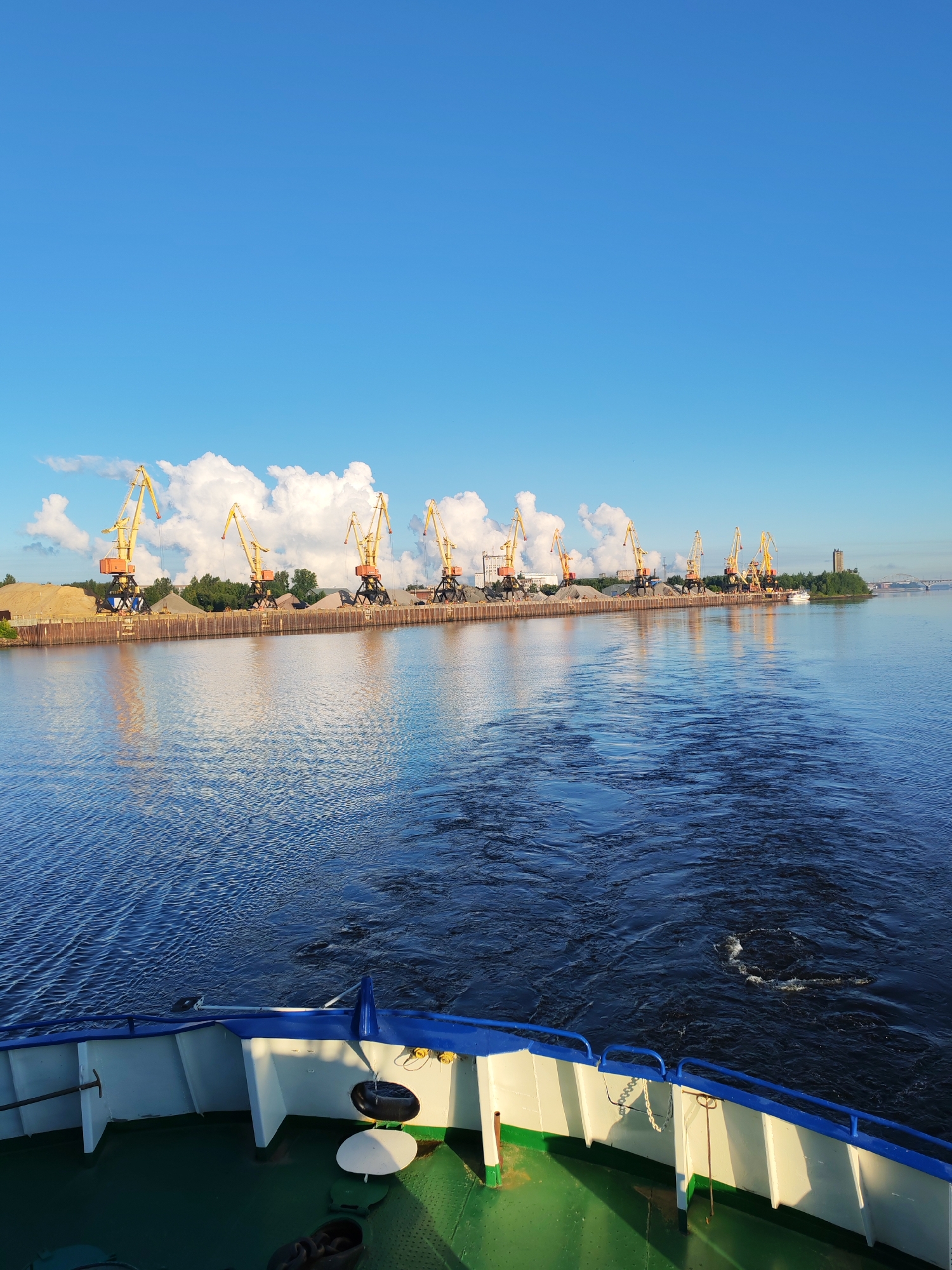 A wide shot captures a body of water with a cargo ship's bow in the foreground. In the middle ground, a line of yellow construction cranes stands against a backdrop of piled earth and trees. The background is a bright blue sky with scattered white clouds. The water shows ripples and a wake, indicating movement. The ship's deck, painted green, has white and blue railings and various metal fixtures. The cranes are industrial and appear to be in a port or industrial area.