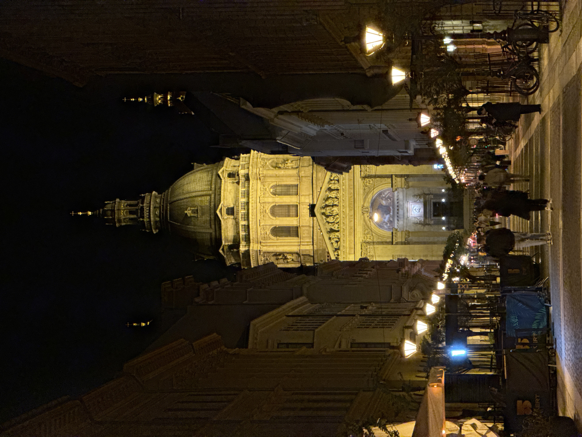 This night-time image captures a grand architectural landmark, likely St. Stephen's Basilica in Budapest, Hungary, beautifully illuminated against the dark sky. The basilica features a prominent dome and multiple spires, with intricate details highlighted by warm, golden lights.
In front of the building, a bustling street or pedestrian plaza is alive with activity. Numerous warm-toned streetlights line the thoroughfare, casting light on people strolling and congregating. Several indistinct figures are visible, some walking, others standing, enjoying the evening atmosphere.
The street is equipped with outdoor seating areas, suggesting cafes or restaurants, and bicycles are parked along railings. Decorative lights are strung overhead, and temporary structures or event setups, possibly for market stalls or outdoor dining, are present. On a dark banner in the lower right, the text "ERT" is visible, and on a blue-patterned covering, "IXI" can be seen. The overall scene depicts a vibrant and well-lit urban environment at night.