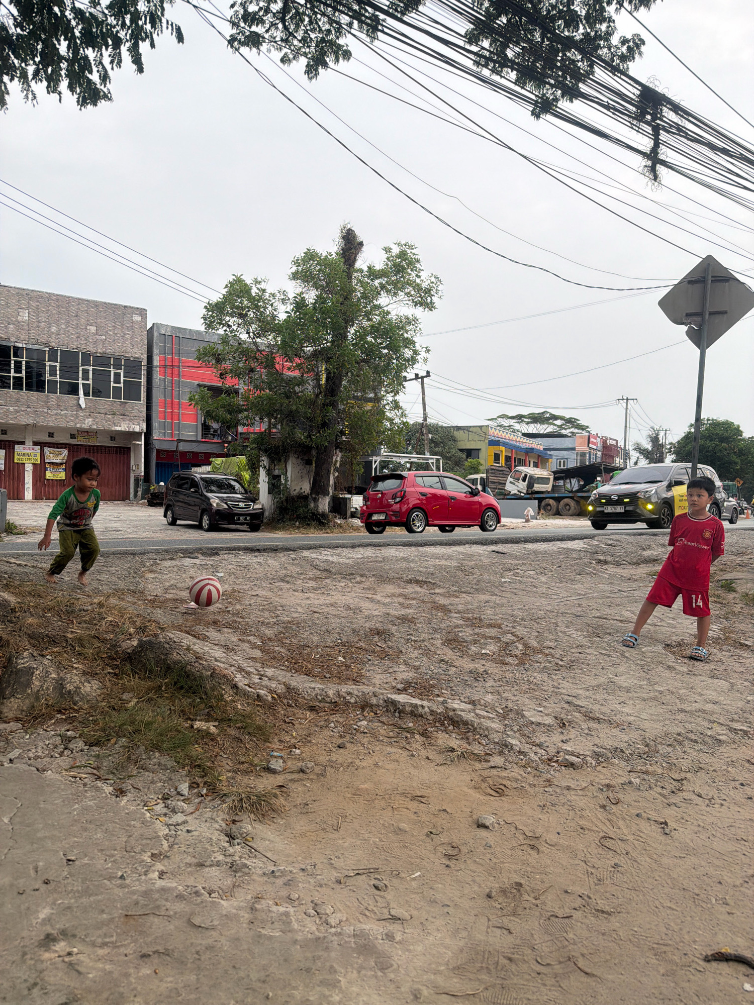 Two young boys are playing soccer on a dirt field next to a paved road in Balikpapan, Indonesia. The boy on the left is wearing a green shirt and pants, while the boy on the right is wearing a red soccer jersey and shorts. A red and white soccer ball is on the ground between them. In the background, there are several buildings and cars on the road. The sky is overcast.