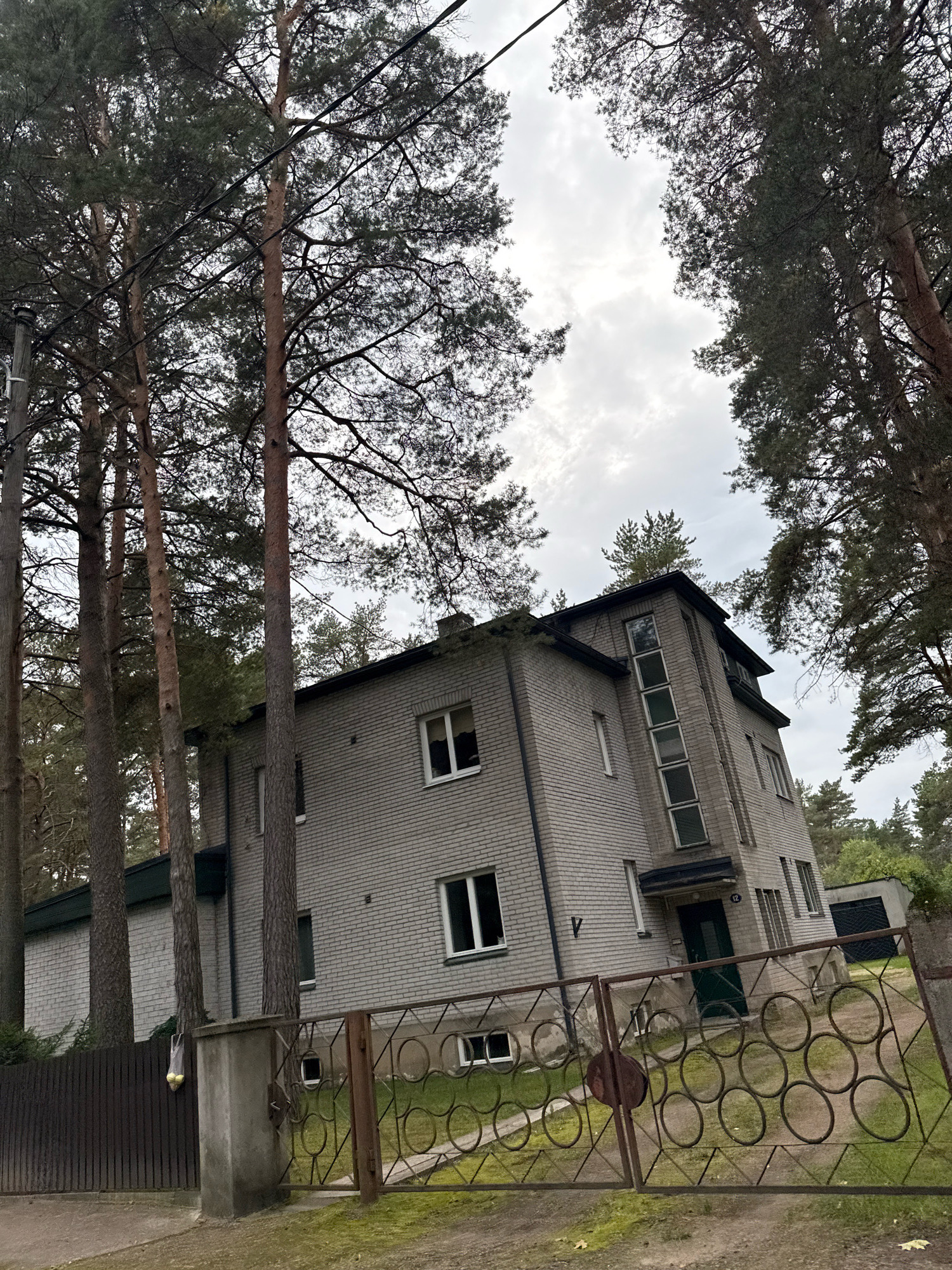 The image depicts a multi-story brick building in a wooded area, likely in Tallinn, Estonia. Tall pine trees surround the property, with power lines visible overhead. The building itself is constructed of light-colored bricks and has a dark roof. It features multiple windows, some of which are arranged vertically on a corner section. A dark metal fence with a circular decorative pattern encloses part of the property, and a wooden fence is also visible. A concrete pillar stands near the entrance. A green door is set into the building, and a small garage-like structure is present in the background. The sky is overcast, suggesting a cloudy day.