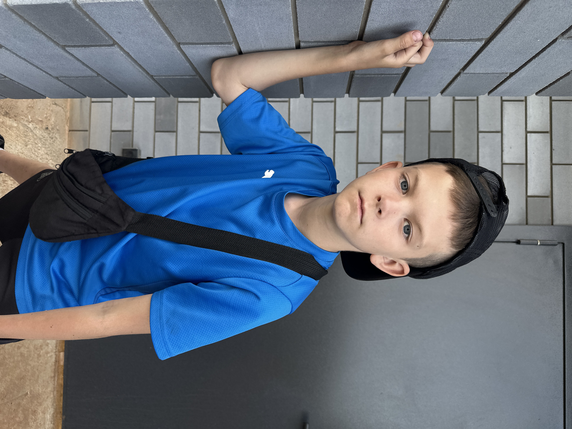 A young male, possibly a teenager, is positioned near a gray brick wall, his hand touching it. He's wearing a blue short-sleeved athletic shirt with a small white logo, black shorts, and a black baseball cap tilted slightly. A black fanny pack is slung across his chest. The setting appears to be outdoors, possibly in the city of Прикамский, Russia, based on the prompt's context. He is looking directly at the camera. There is a grey door next to him.