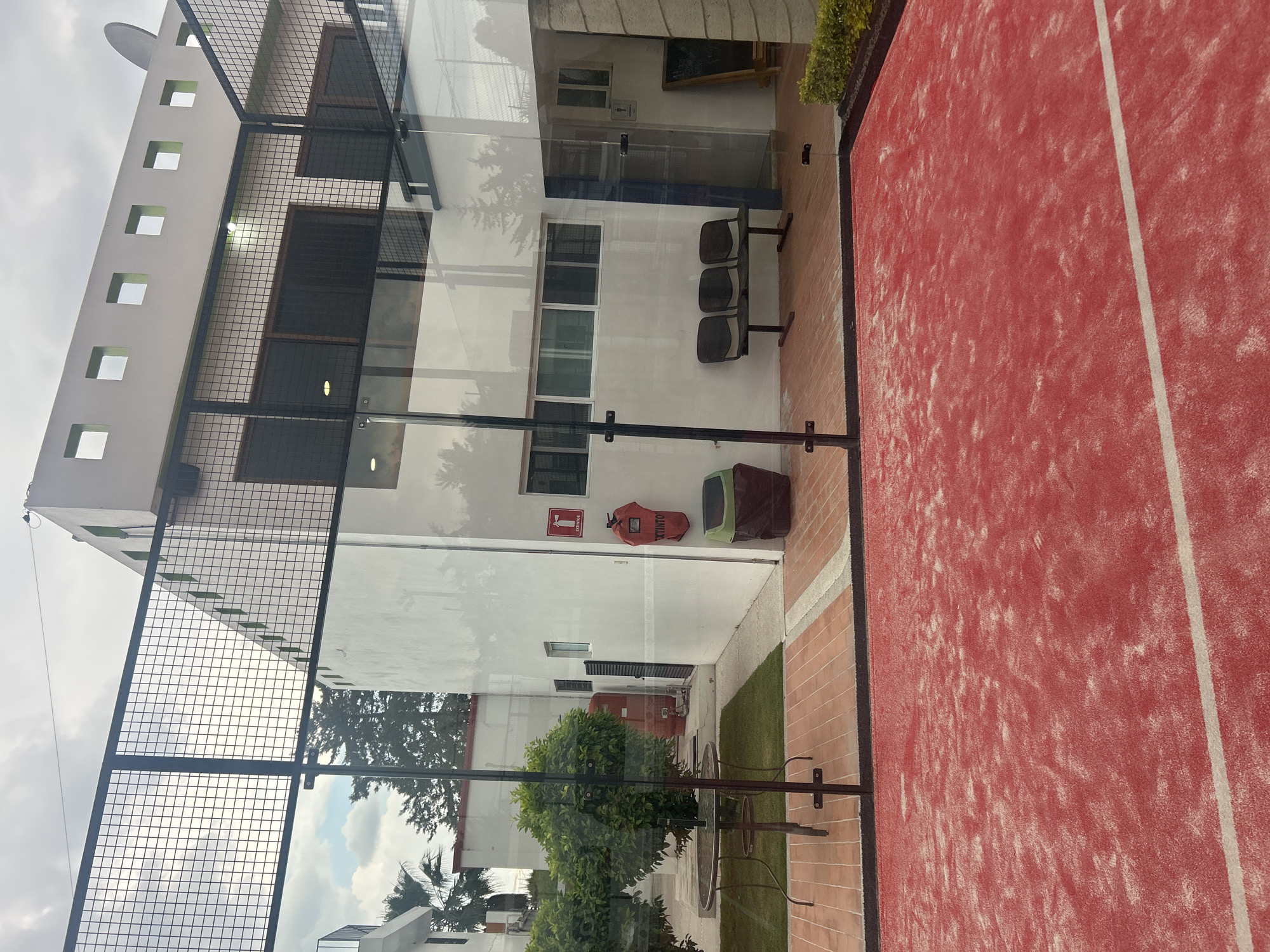 The image shows a paddle tennis court in Puebla, Mexico. The court is red with white lines. Adjacent to the court, there is a building with a white facade, metal mesh fencing, and glass panels. The building has several windows and a satellite dish on the roof. There are three chairs near the building and a fire extinguisher, along with other safety equipment on the wall. A small garden area is visible with trees and a table and chairs. The sky is cloudy.