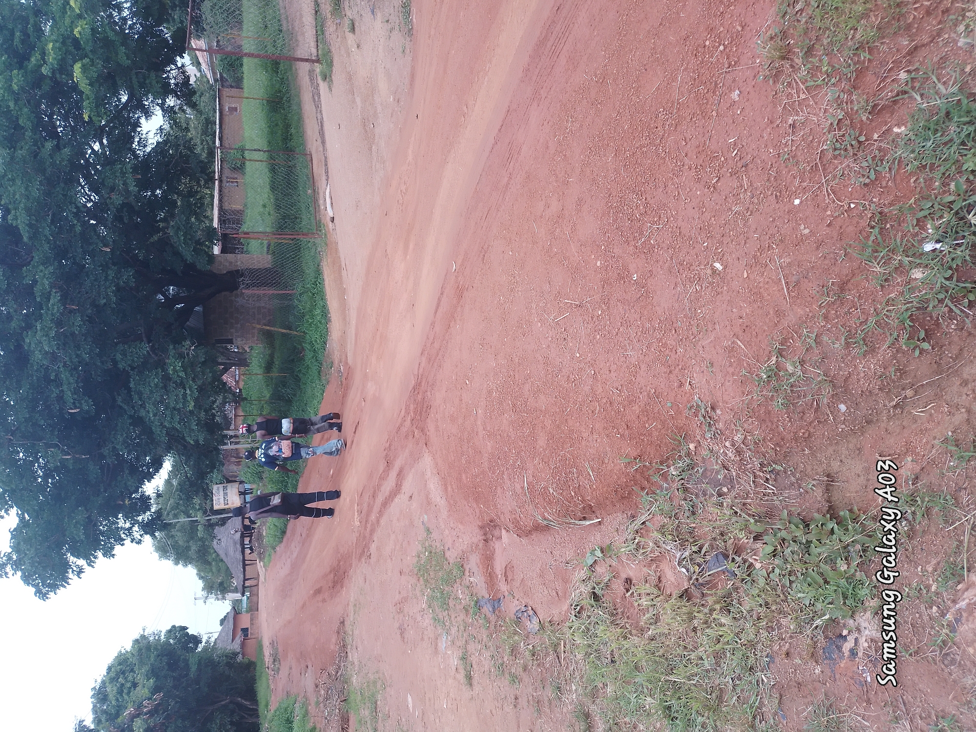 Three young males walk down a dusty, reddish-brown dirt road in an outdoor, rural setting. The males are dressed casually and are walking away from the camera. One male, in the center, is wearing a blue shirt with a backpack, and the other two are wearing dark shirts and pants. The road is flanked by sparse green vegetation and some scattered debris. In the background, there are traditional-looking buildings with thatched roofs, a large tree, and a chain-link fence. The sky is overcast. The location is identified as Unknown, Unknown.