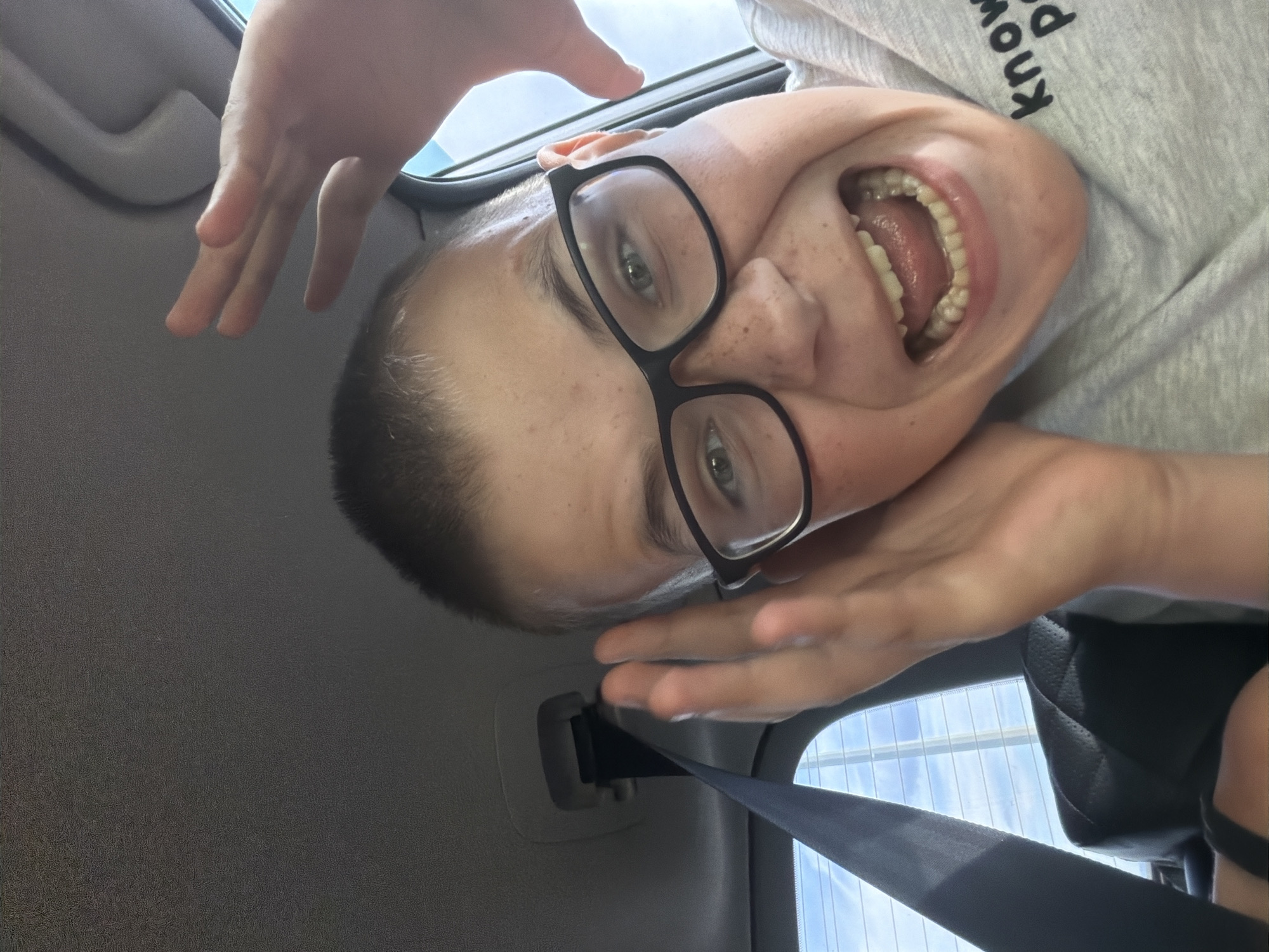 The image is a close-up, low-angle selfie taken from inside a vehicle. A young person with short brown hair, wearing black-rimmed glasses and a grey t-shirt with the partial text "know P", is making a surprised or excited facial expression with their mouth wide open and hands near their ears and face. They are seated in a car, with a grey car interior visible, including a seatbelt and the car's headliner. The background through the rear window shows a bright sky with some clouds and what appears to be a building or structure with horizontal lines. The image does not contain any discernible information about the city of Новое Маврино, Russia.