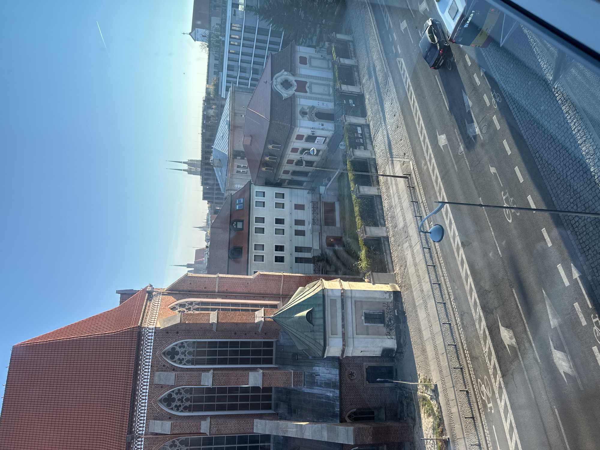 The image depicts a cityscape of Wrocław, Poland, viewed from an elevated position, likely a window. The foreground features the tiled red roof and Gothic architecture of the Cathedral of St. John the Baptist. Beyond the cathedral, various buildings line the street, including residential structures with white and red facades and a modern office building. A street with bicycle lanes and road markings runs adjacent to the buildings. A dark-colored car is seen on the street. In the distance, the spires of other churches are visible against the clear, blue sky. The light suggests it's daytime.