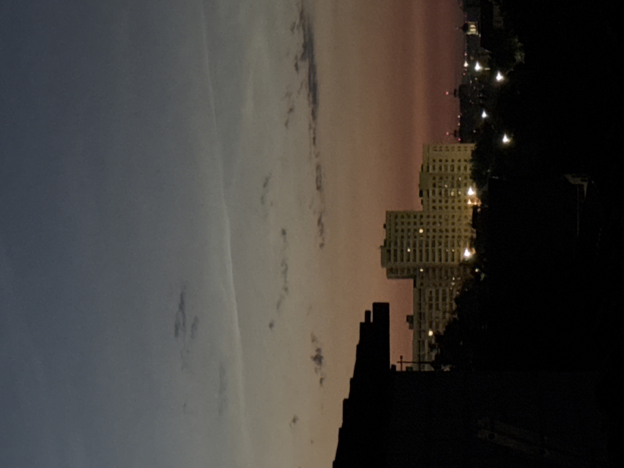 The image captures a twilight view of Novaya Usman', Russia, showcasing an urban landscape with buildings and the sky. In the foreground, a silhouetted structure, likely a roof or building section, juts in from the left side, obscuring much of the immediate surroundings. Beyond this, a cluster of apartment buildings is visible, illuminated by numerous lit windows, suggesting residential occupancy. These buildings are of varying heights and architectural styles, with the most prominent being a taller, lighter-colored structure with a grid-like pattern of windows.
Further in the distance, other buildings and city lights are discernible, hinting at the broader extent of the urban area. The sky transitions from a deep blue or grey at the top to a warm, reddish-orange hue closer to the horizon, characteristic of sunset or dawn. Wispy clouds are scattered across the sky, some appearing as faint streaks. The overall atmosphere is one of a quiet evening or early morning in a residential part of the city. There are no visible people in the frame. The scene is static, devoid of any discernible action or interaction. The lighting is dim, with artificial lights from the buildings contrasting with the fading natural light.
