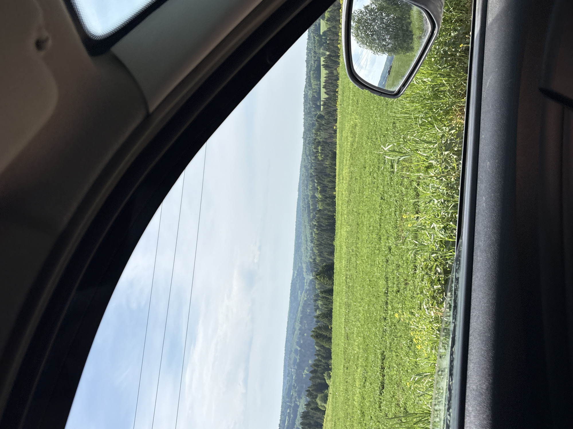 The image shows a view from inside a car. Outside the car window is a green field or meadow. A line of trees or a forest appears in the distance. The car's side mirror is visible, reflecting a view of the surrounding landscape. The sky is light blue with some clouds. The image does not contain enough information to verify its location in Khokhlovka, Russia, or any details about people, specific objects, actions or interactions.