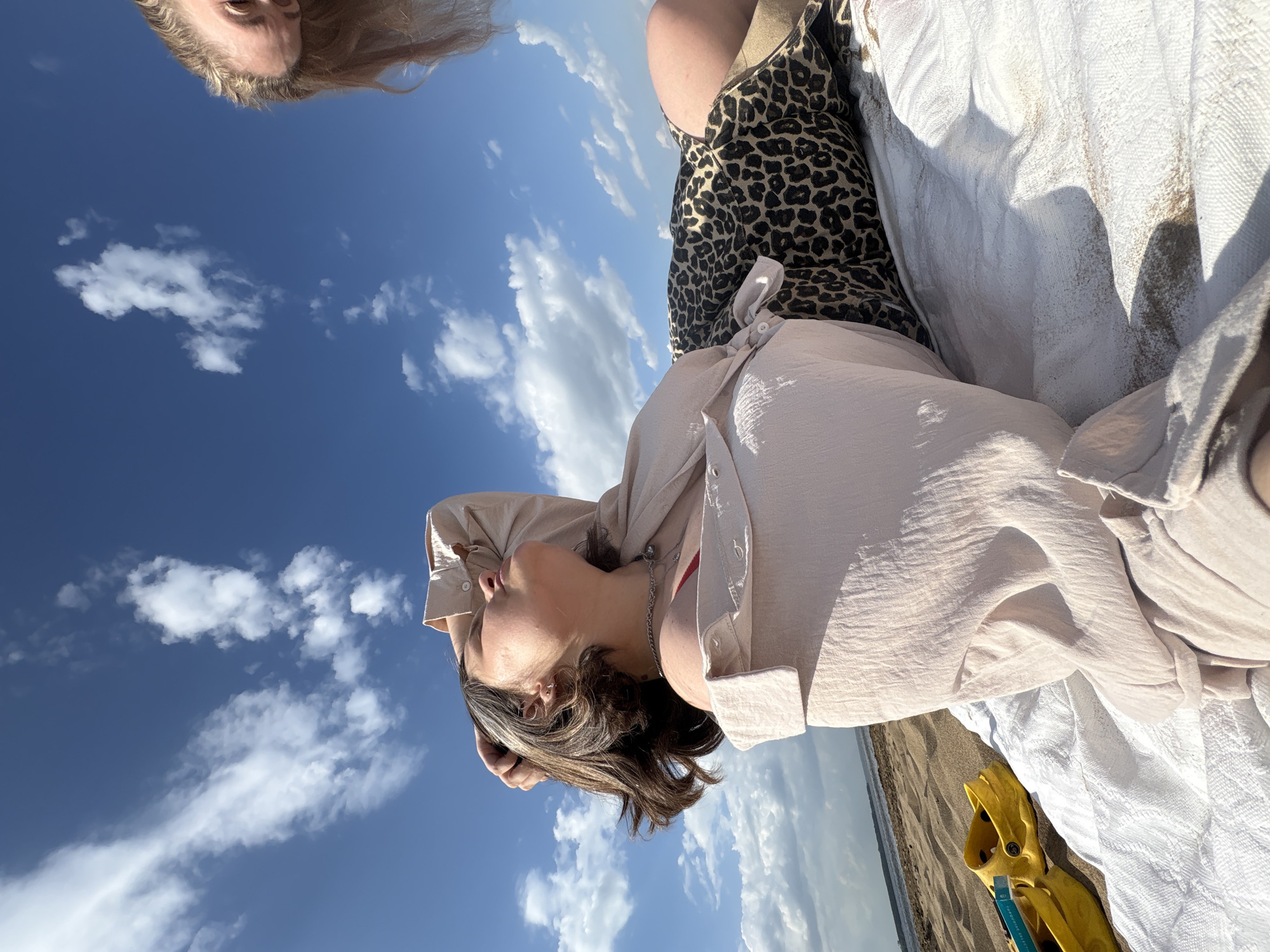 The image depicts two women at a beach in Chaikovsky, Russia. One woman is lying on a white blanket, wearing a tan-colored shirt and a leopard-print dress. Her head is angled towards the sky, and she is looking up. Next to her, there are yellow sandals and a book. The other woman is partially visible at the top of the frame. The background shows a blue sky with white clouds, the beach, and the sea.
