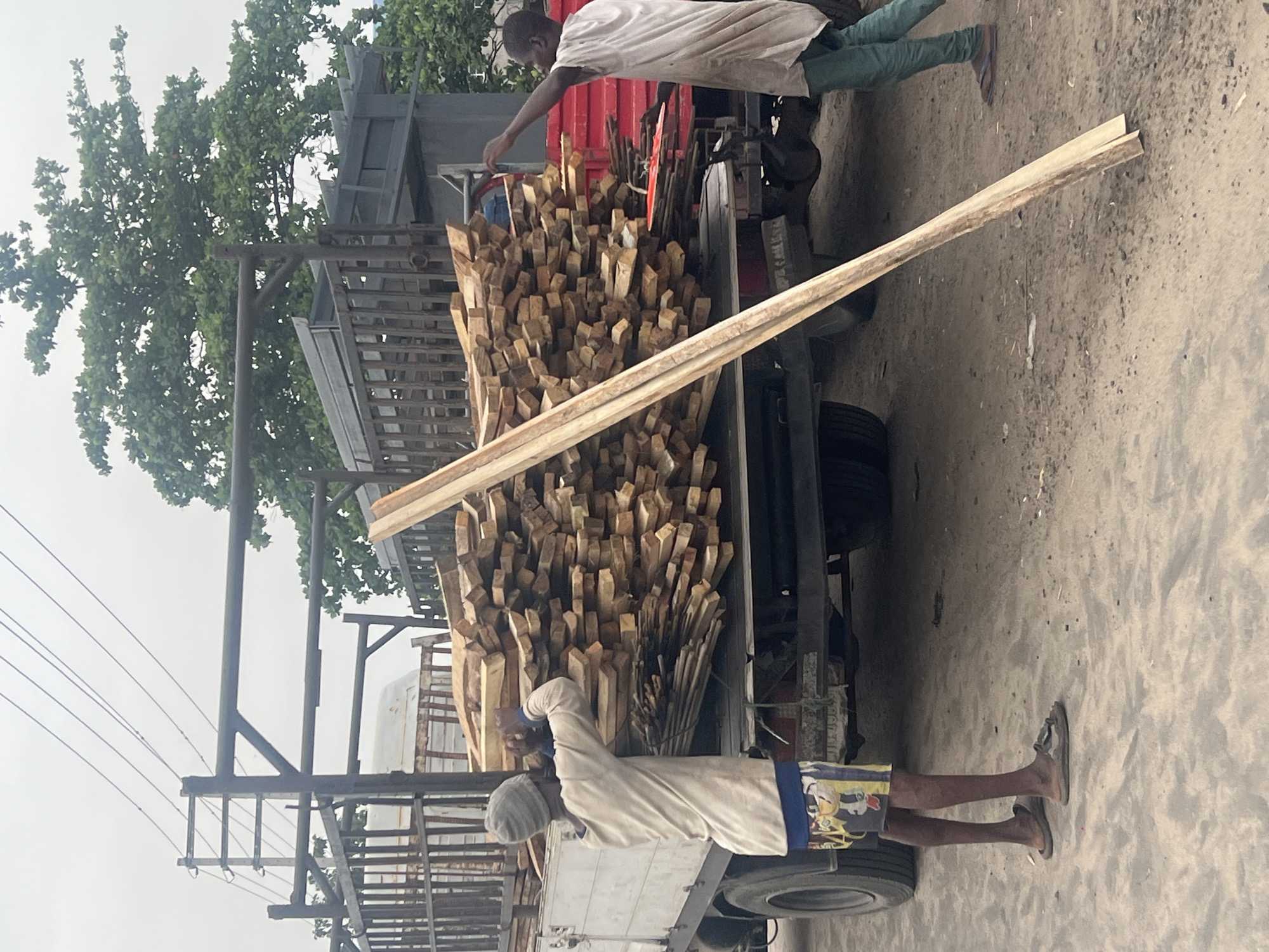 Two men are loading a truck with wooden planks. The truck is packed with an abundance of short, square-cut pieces of wood, mostly a light tan color. The man on the right is standing on the truck bed, wearing a light-colored, long-sleeved shirt and light green pants. He is bending down, seemingly adjusting the load. The man on the left is on the ground, wearing a light-colored shirt and patterned shorts. He appears to be holding a long wooden plank that is being loaded onto the truck. The ground is sandy. There are power lines visible in the background. The setting appears to be a street in Lagos, Nigeria.