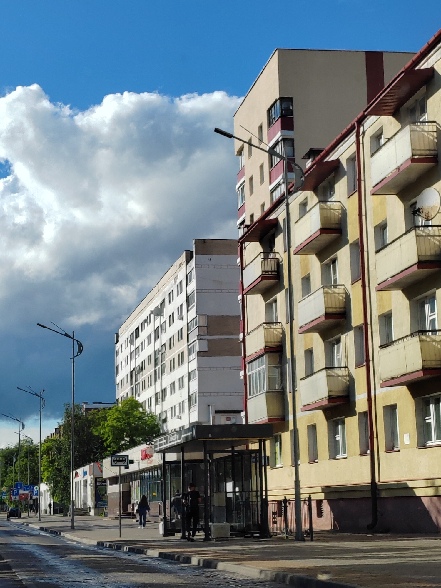 A street scene in an urban environment features multiple apartment buildings under a partly cloudy blue sky. In the foreground, a crosswalk crosses a paved street, and to the right, a bus stop shelter is visible. Several people are present, including two individuals standing at the bus stop, one of whom appears to be a security guard in uniform. Further down the sidewalk, a woman walks away from the bus stop, and other pedestrians are seen in the distance. On the left side of the street, traffic lights and road signs are present. The buildings are multi-story residential structures with visible balconies and windows. The overall scene depicts a typical day in a city.