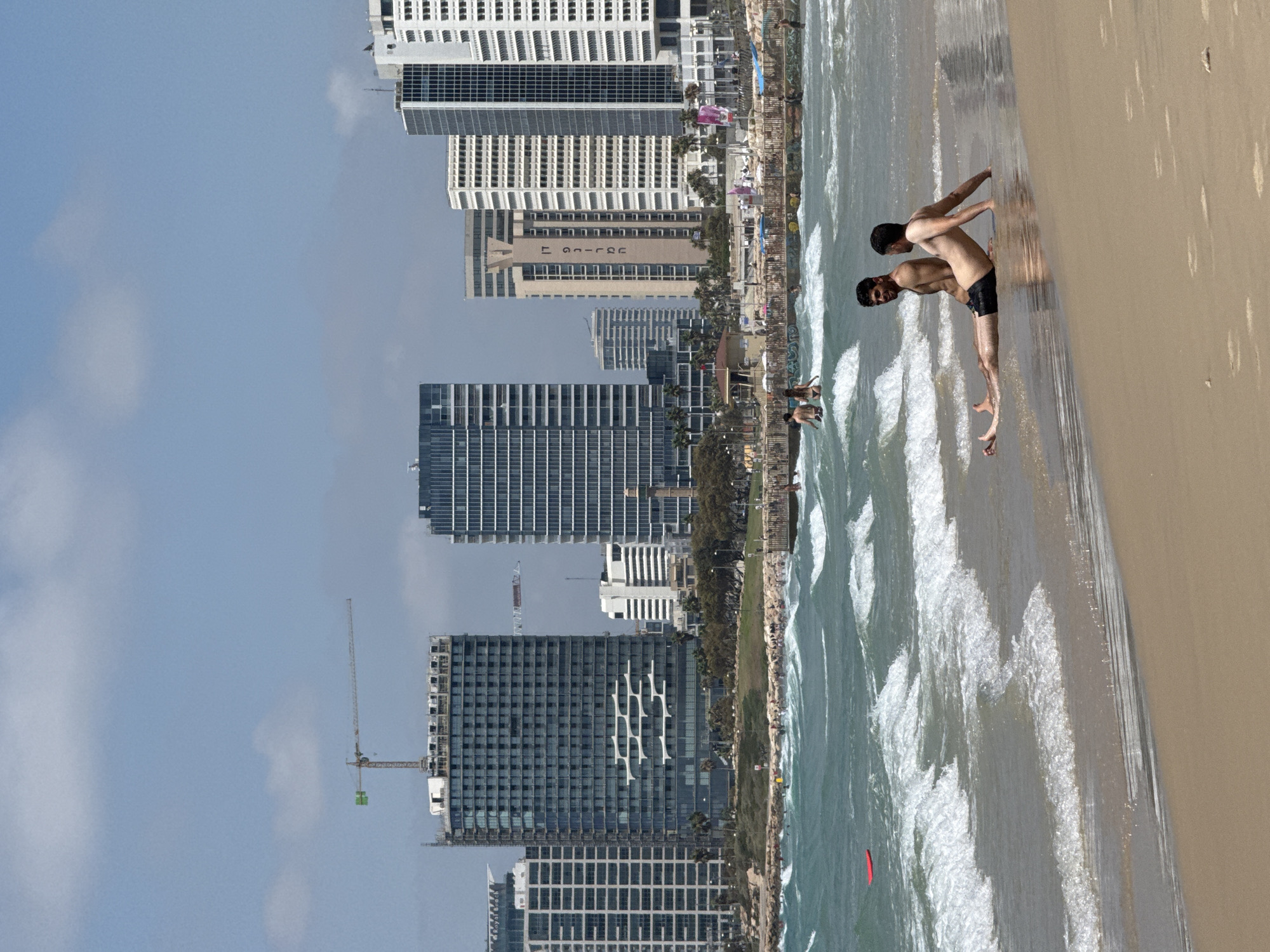 The image captures a sunny day at Tel Aviv's coastline, likely along the Mediterranean Sea. In the foreground, two men are sitting on the wet sand at the edge of the water, one leaning back and the other facing forward. Gentle waves are washing over their legs. The sea is a vibrant turquoise, with white foam cresting on the waves.
Beyond the beach, a bustling promenade is visible, lined with people and some beach structures like umbrellas and lifeguard stands. In the mid-ground, a rocky breakwater separates the beach from the water.
The background is dominated by the distinctive skyline of Tel Aviv, featuring a mix of modern high-rise buildings. One prominent building in the center has a unique architectural design with horizontal white elements on its facade. Further in the distance, construction cranes indicate ongoing development in the city. The sky is clear blue with a few scattered white clouds. The overall scene depicts a lively day at a popular urban beach.