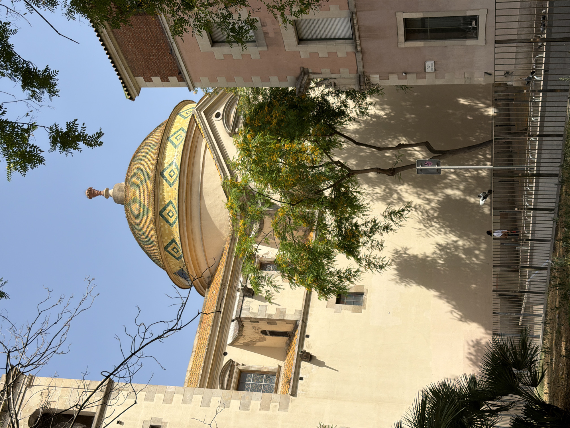 The image captures a low-angle view of a building's exterior in Barcelona, Spain. A prominent, colorful dome, tiled with green and yellow diamond patterns, is a central subject. Adjacent to the dome is a beige-colored building with a red-brick section on the upper left. Several windows with metal gratings are visible on the buildings. Green trees with small yellow flowers partially obscure the view of the building and dome, casting shadows on the facade. In the background, a metal fence runs along a walkway. A street sign with the text "PLAÇA CASTELLÀ" is affixed to the wall. The scene appears to be set during the daytime, under clear, blue skies. A few birds are perched on the fence and one is in flight. A person can be seen in the background near the fence. The overall impression is one of a bright, sunny day in an urban setting.