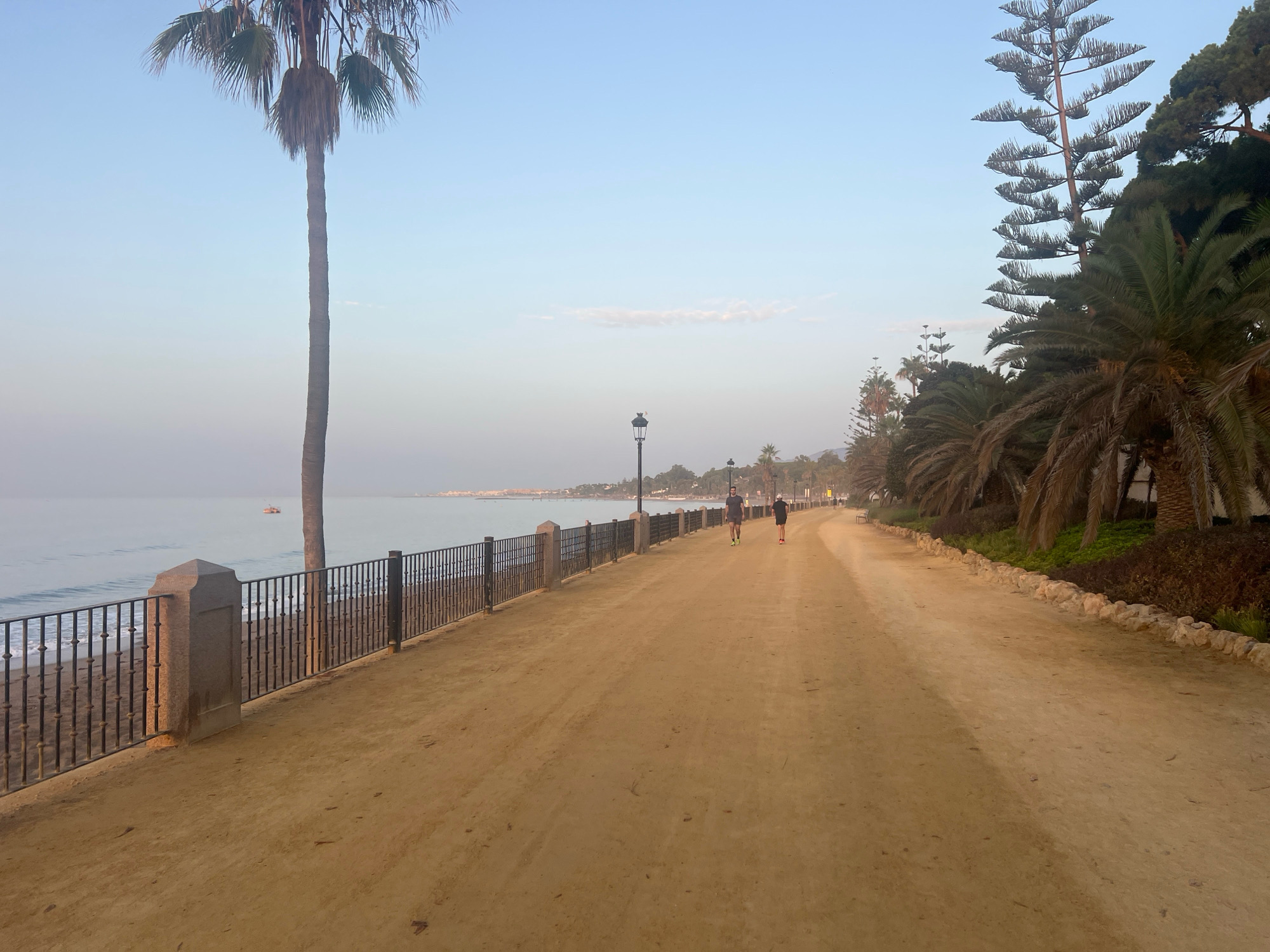 The image shows a dirt path along the coast of Marbella, Spain, with the sea on the left and vegetation on the right. Two individuals are jogging on the path in the distance. The path is bordered by a metal fence and stone pillars on the left, and a line of rocks and palm trees on the right. A tall palm tree is visible in the foreground on the left, and other palm trees and a large pine tree are seen further down the path. The sky is clear and pale blue, suggesting it is early morning or late afternoon. The sea is calm with gentle waves breaking on the shore. A small boat is visible in the water on the left. Streetlights are spaced along the path.