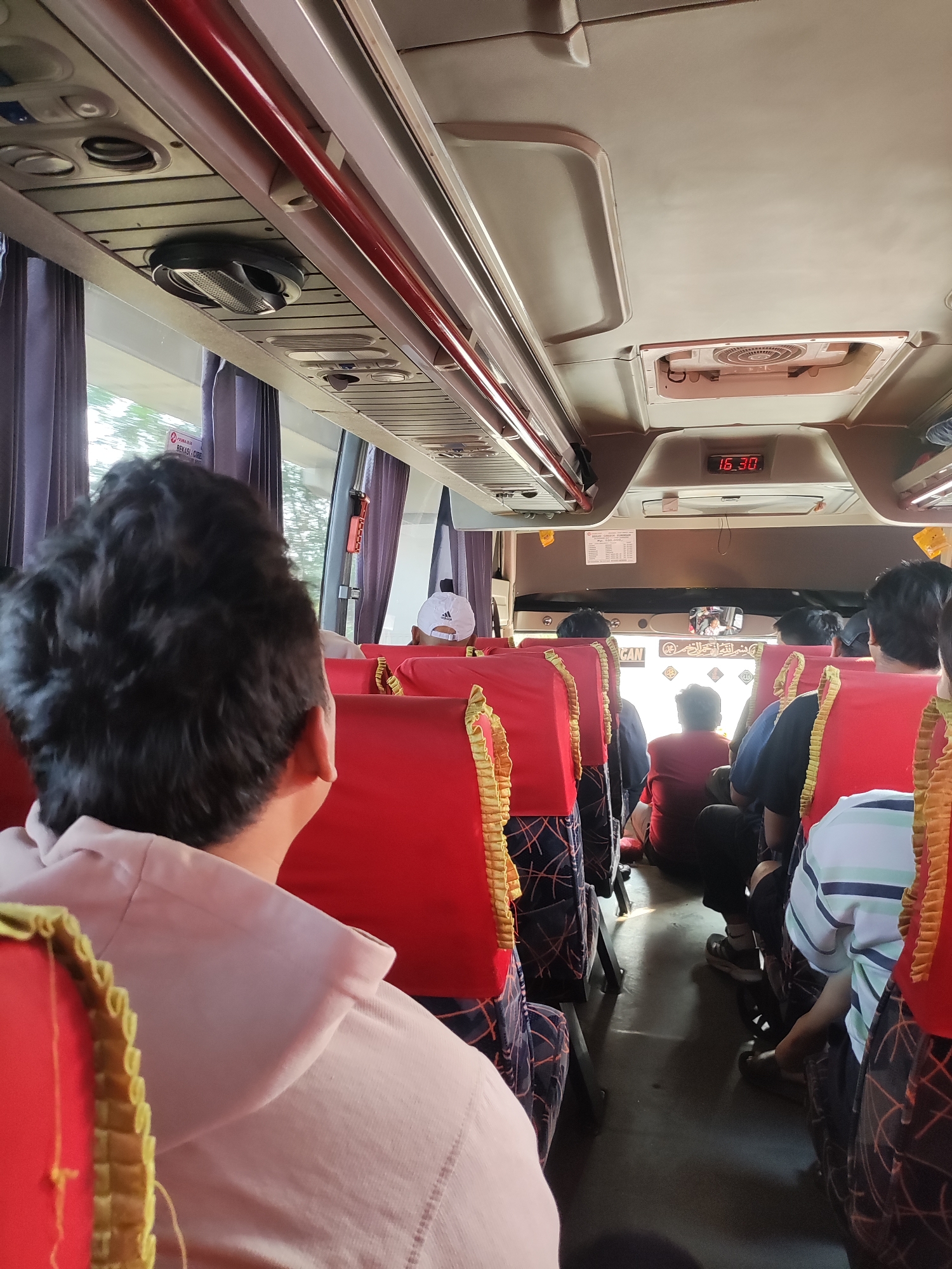 The image shows the interior of a bus in Karawang, Indonesia, with passengers seated on red seats. In the foreground, the back of a person's head with dark, curly hair is visible, wearing a light pink shirt. The bus aisle is visible, with seats on either side. There is a digital clock on the ceiling displaying "16:30". Luggage racks and air vents are also visible on the ceiling. Outside the window, greenery is visible. The overall scene depicts a typical bus journey in Indonesia.