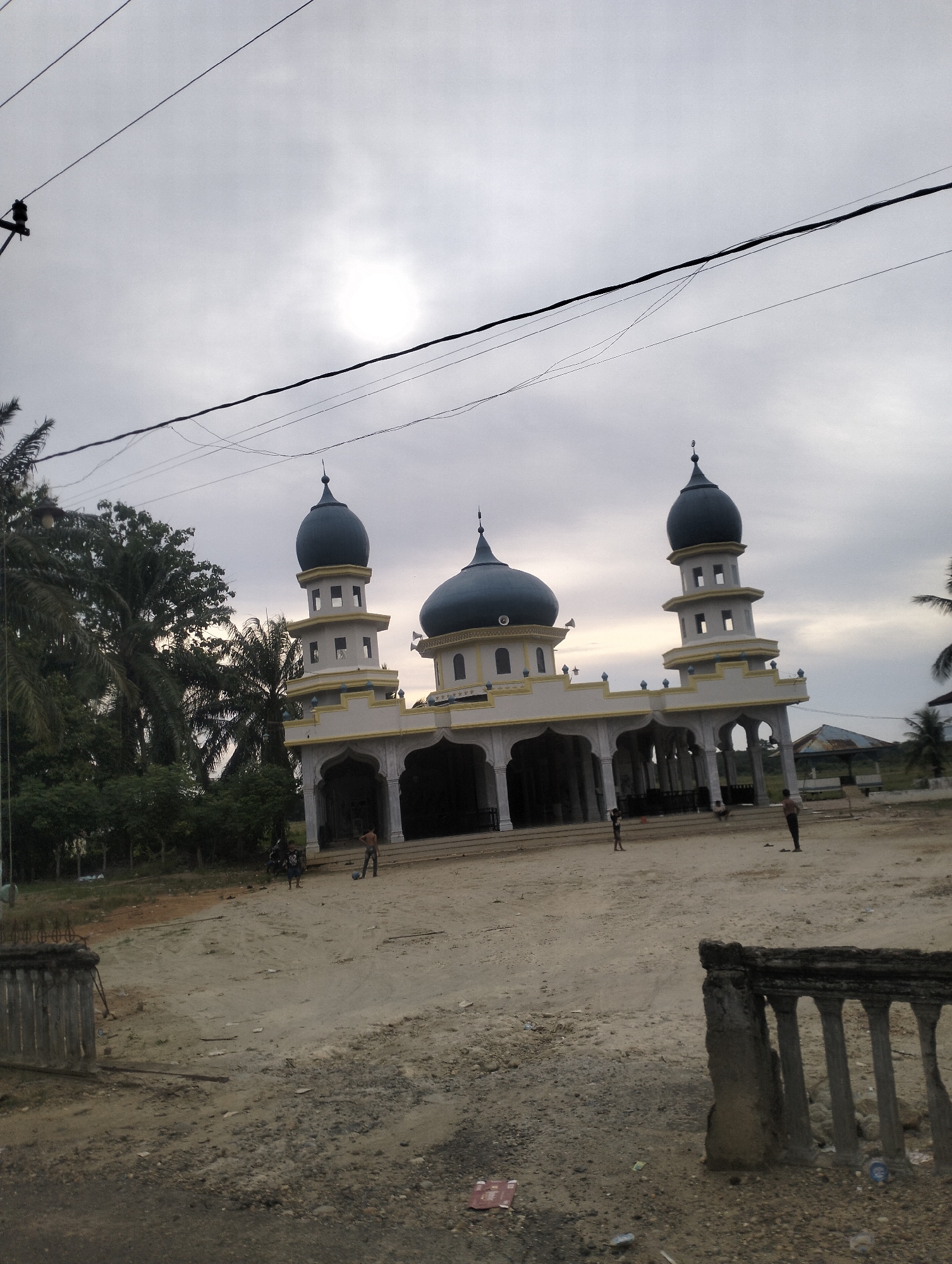 A mosque with three domes and two minarets stands under a cloudy sky. The mosque is light-colored with yellow accents. In front of the mosque, there is a sandy courtyard where several people are present, some of whom appear to be children playing. Tall palm trees and other vegetation surround the mosque. Power lines are visible in the foreground and across the sky. The ground in the foreground is a mix of dirt and gravel, with a low-lying fence or wall on the left side. Another similar structure, possibly a decorative fence, is on the right side of the foreground. The overall impression is a daytime scene in a rural or semi-rural setting.