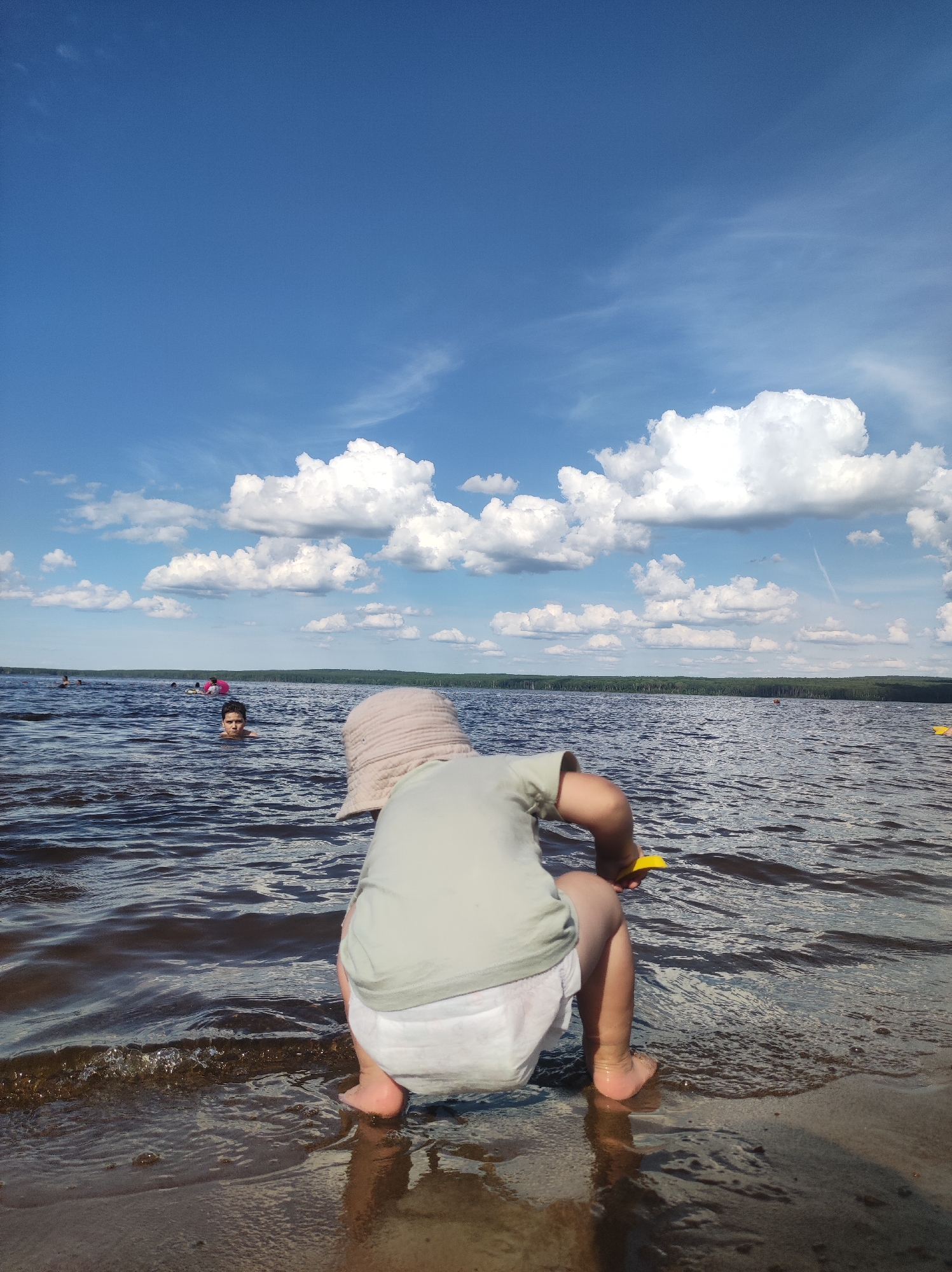 An outdoor, eye-level shot captures a young child from the rear, crouching at the edge of a body of water. The child wears a light-colored hat, a pale green t-shirt, and a white diaper. They are holding a yellow toy in their right hand and have their left foot submerged in the shallow water. The water's surface is rippled, with small waves lapping at the sandy shore.
In the background, the lake stretches towards a tree-lined horizon under a bright blue sky filled with fluffy white clouds. Several other individuals are visible in the water, some swimming and others with inflatable rings, suggesting a public recreational area. The scene is likely set at a beach or lakefront in Chaikovsky, Russia, given the context of similar images from the area. The overall atmosphere is one of a sunny day spent enjoying outdoor activities by the water.