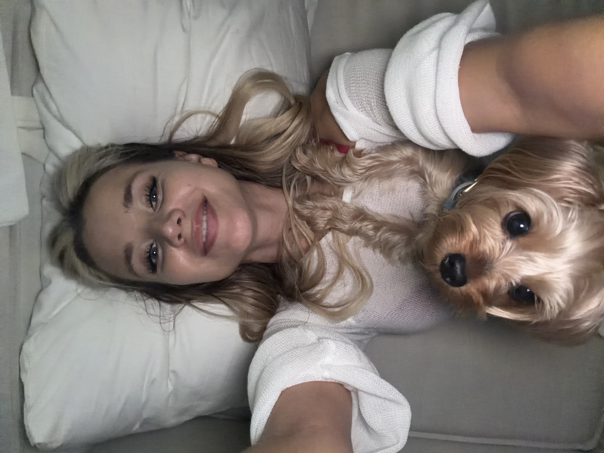 The media file is a selfie of a young woman and a small, light brown dog. The woman has long, blonde, wavy hair and is smiling at the camera. She is wearing a white, textured sweater. The dog, which appears to be a Yorkshire Terrier, is looking directly at the camera with its head tilted. It is wearing a light blue collar. They are both lying down, possibly on a couch or bed, with white pillows visible in the background. The lighting suggests it might be indoors, possibly in the evening or on a cloudy day, as there are no strong shadows. The overall mood is relaxed and affectionate, with both subjects appearing comfortable and happy. There is no visible text in the image. The setting is likely a domestic environment.